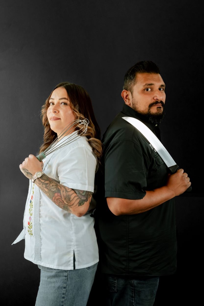 A man and a woman stand back-to-back against a black background. The woman is holding a metal whisk and has tattoos on her arm. The man is holding a large kitchen knife. Both have a confident expression.