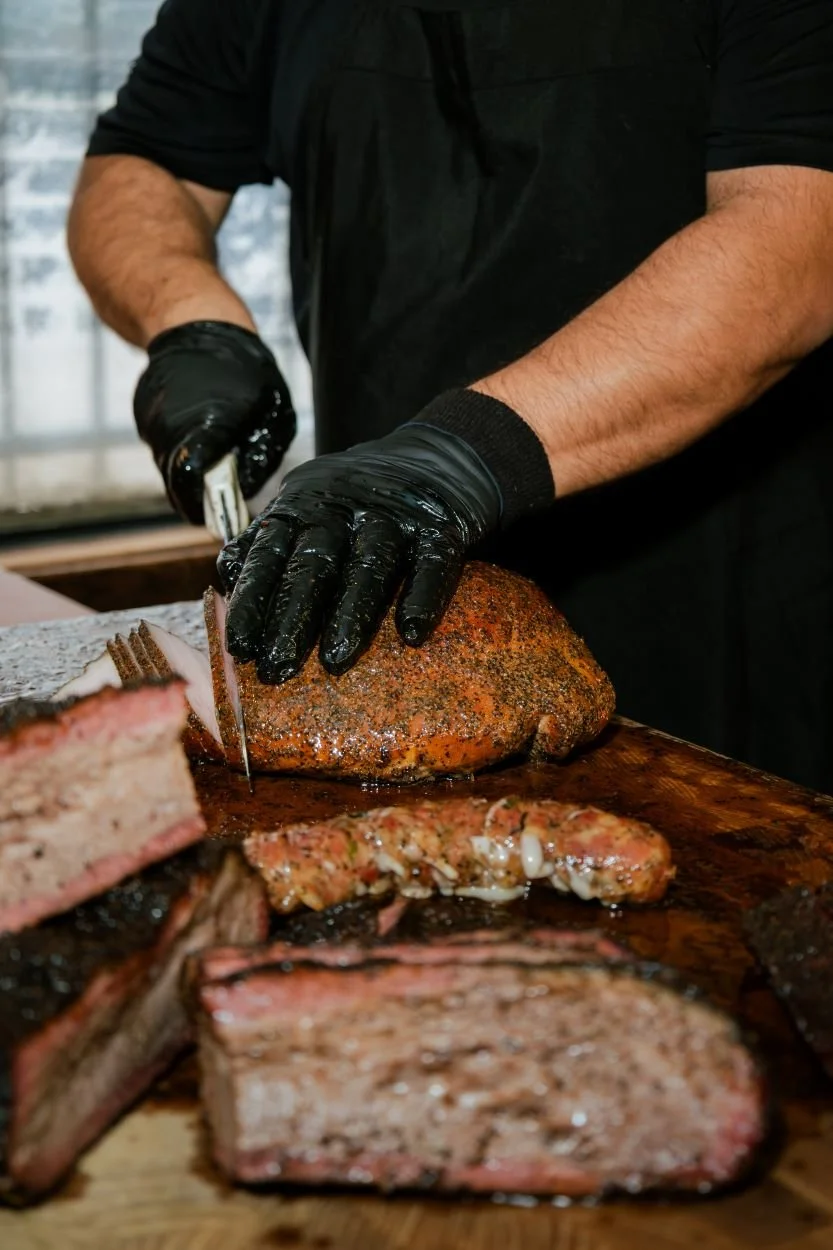 A person wearing black gloves and a black shirt is carving a seasoned, cooked brisket on a wooden cutting board. The scene suggests preparing smoked meat, with other cuts of meat visible in the foreground.