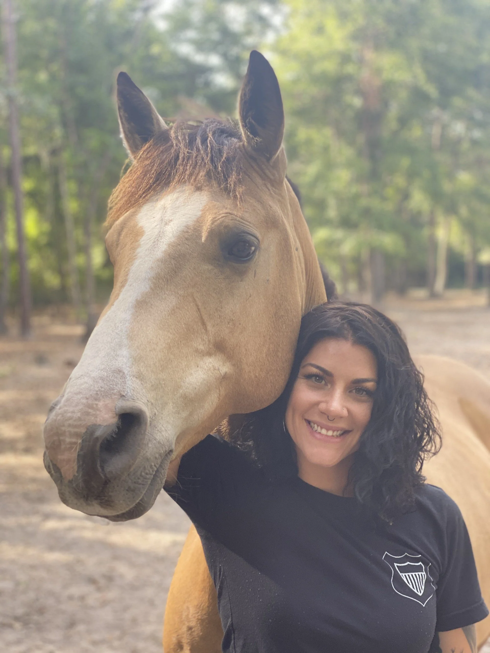 A woman with curly dark hair smiling while taking a selfie with a horse. The horse's head is close to the woman's face, and the background shows a wooded outdoor area.