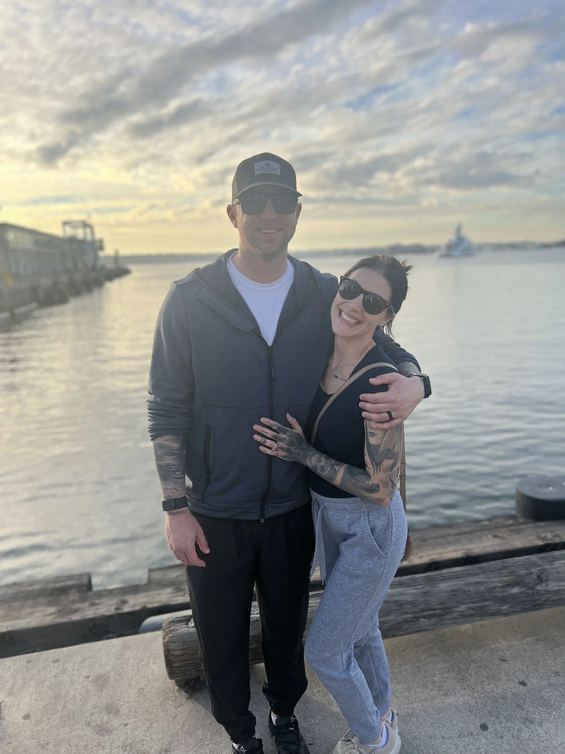 A smiling man and woman hugging at the waterfront during sunset, with boats in the background and a cloudy sky.