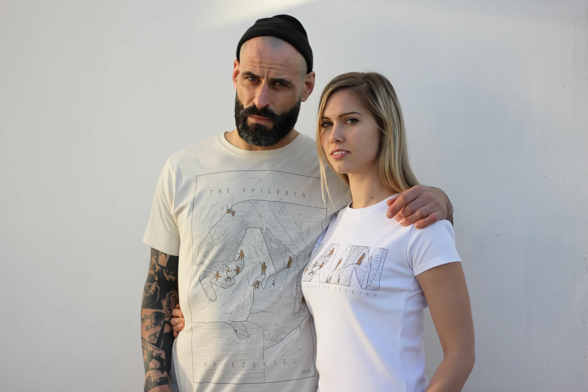 A man with a beard wearing a black beanie and a woman with long blonde hair wearing a white T-shirt, standing close together outdoors in front of a plain white wall, with the man's arm around the woman's shoulder.