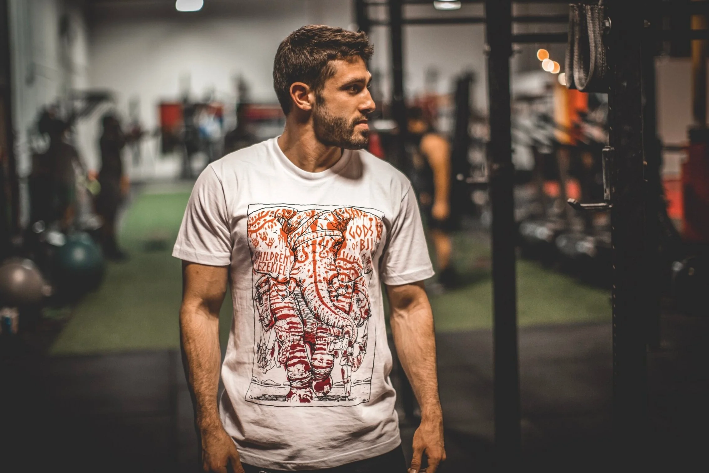 A man with short dark hair and a beard wearing a white graphic t-shirt with a detailed elephant illustration, standing in a gym.
