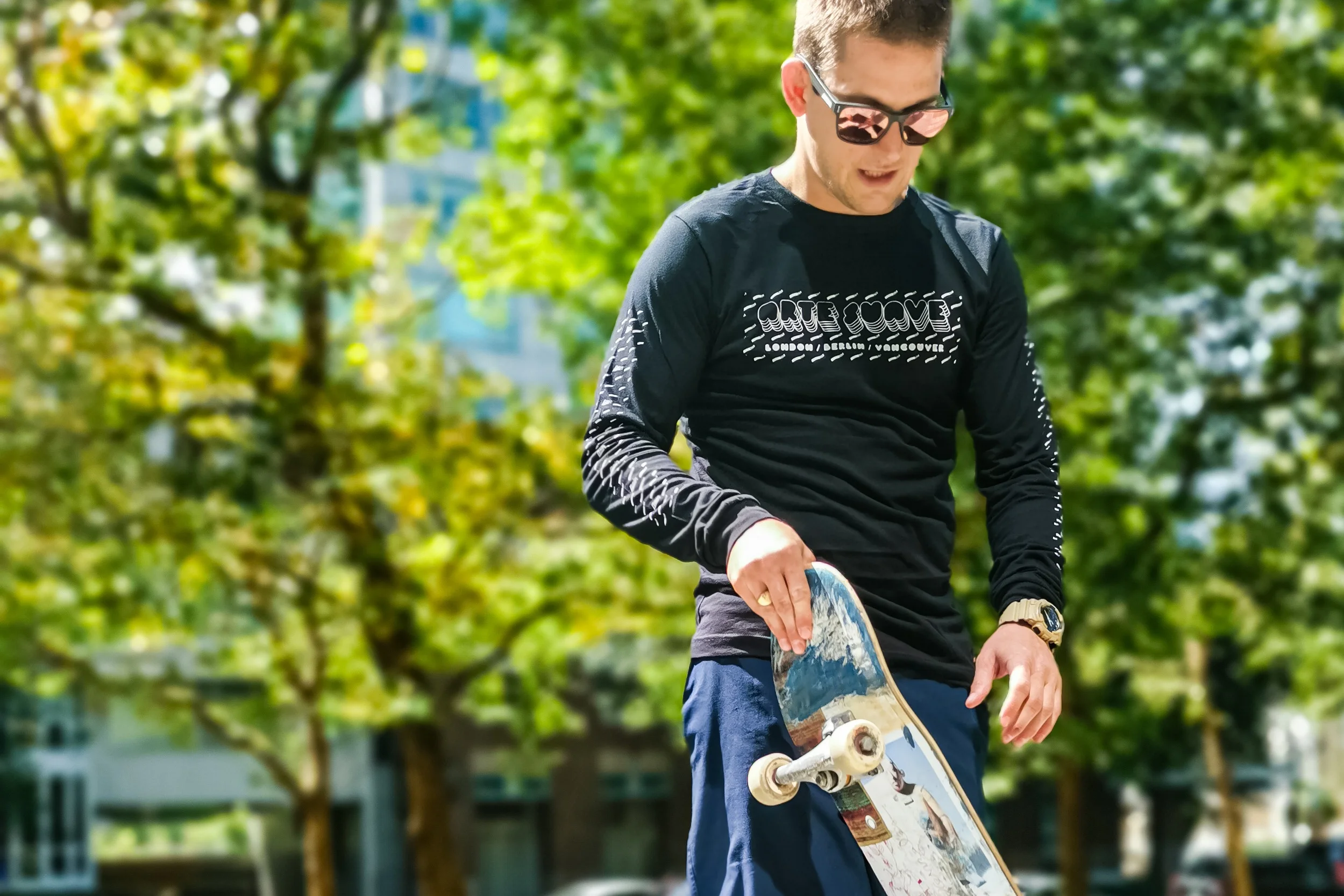 A young man wearing sunglasses and a black long-sleeve shirt with the words "Corte Couvre" on it, holding a skateboard, standing outdoors surrounded by green trees.
