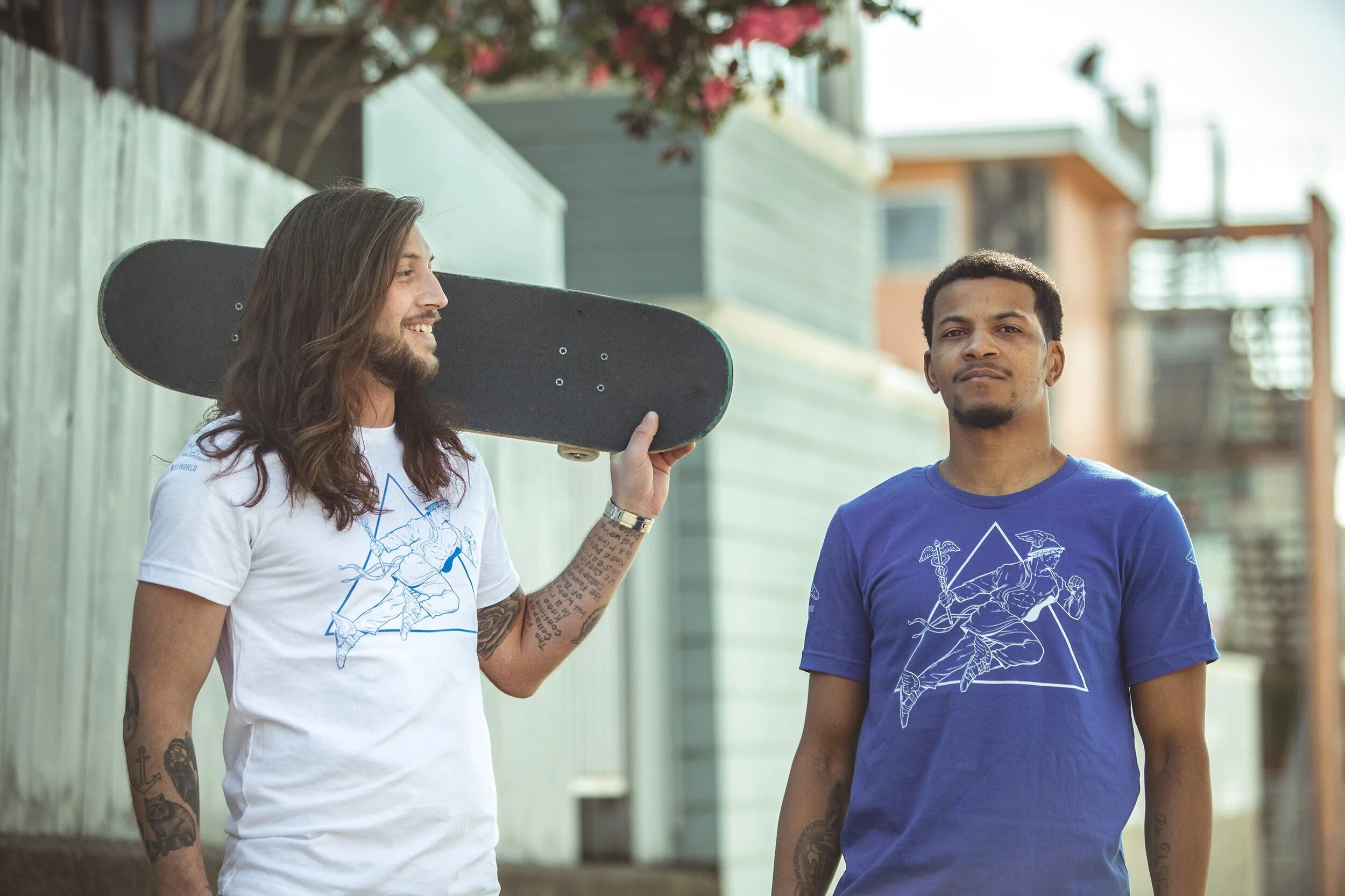 Two men walking outdoors, one with long hair and tattoos holding a skateboard on his shoulder, and the other with short curly hair, both wearing graphic t-shirts, in front of a residential building with wooden fencing and flowering plants.