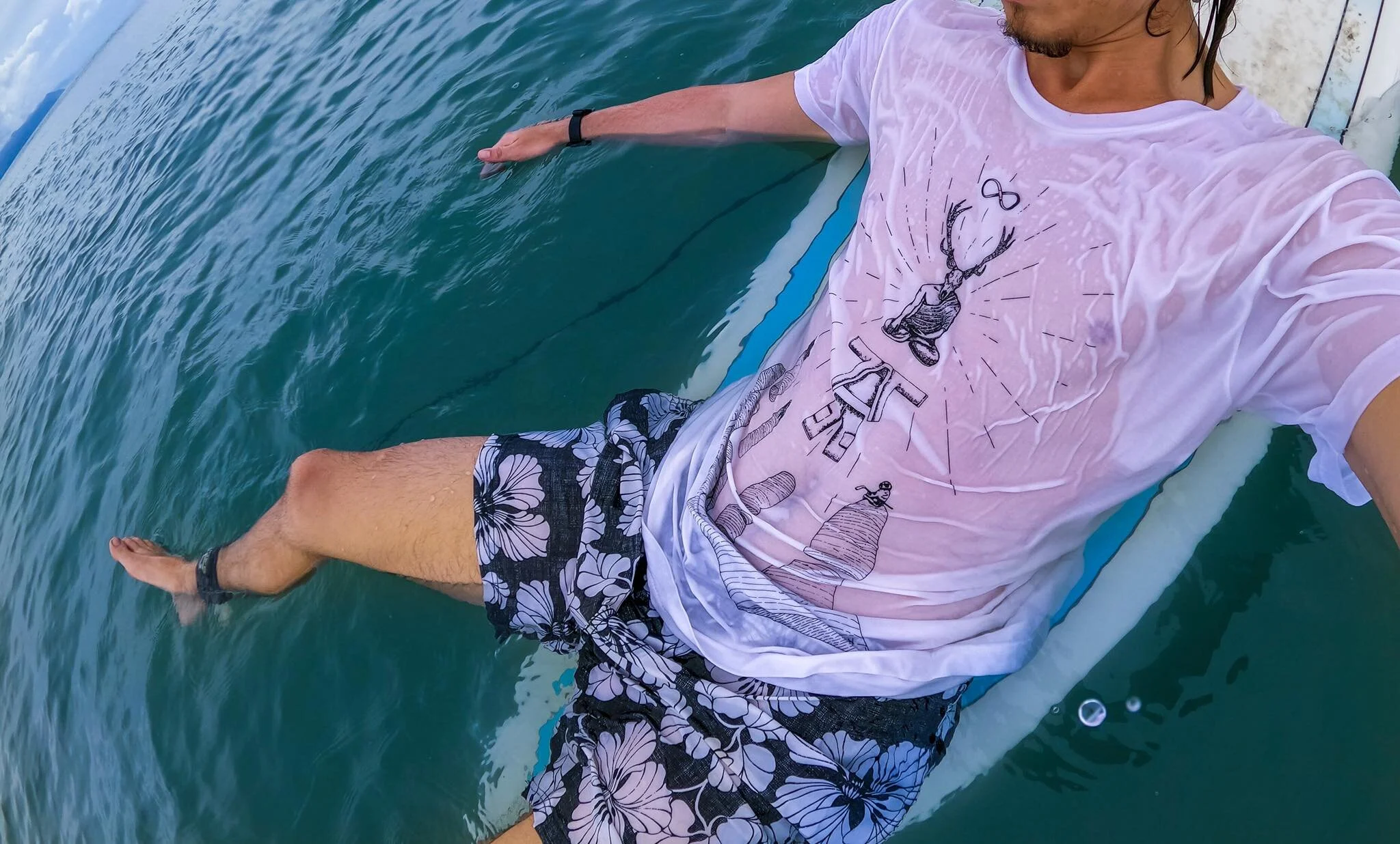 A person on a surfboard in the water, wearing a pink t-shirt with a graphic design and black floral swim trunks.