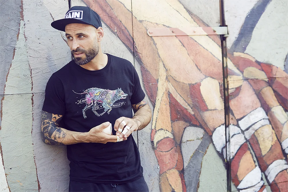 A man with a beard and tattoos wearing a black t-shirt with a colorful wolf graphic and a black cap, standing against a graffiti wall.