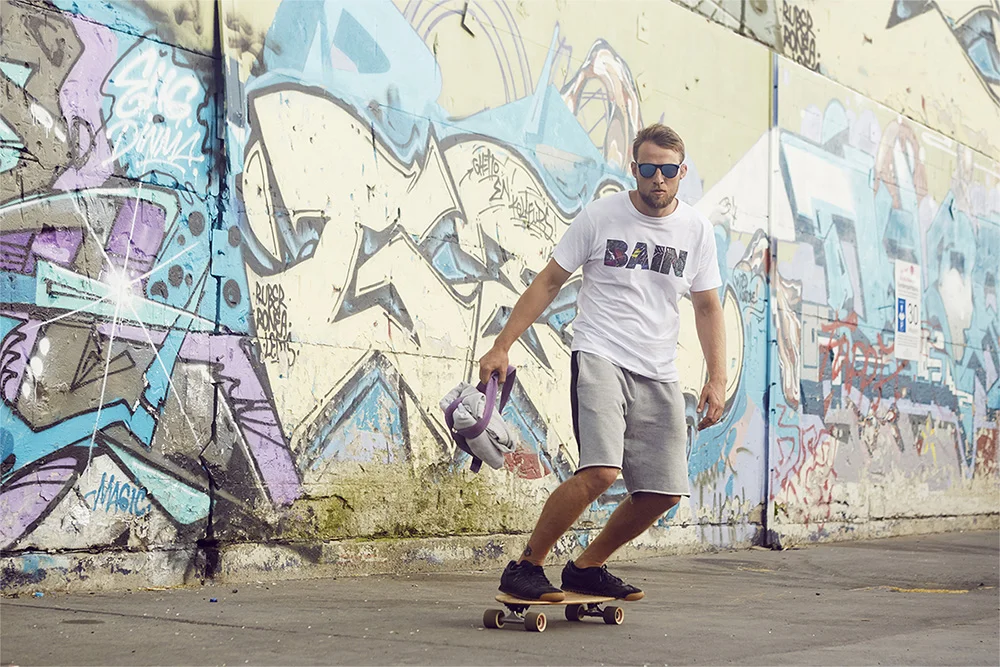 A man skateboarding in front of a colorful graffiti wall, carrying a purple helmet and gray hoodie.