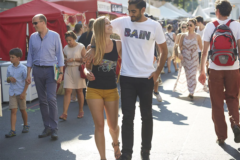 Young couple walking at an outdoor fair or festival, smiling and looking at each other, surrounded by other people and vendor tents.