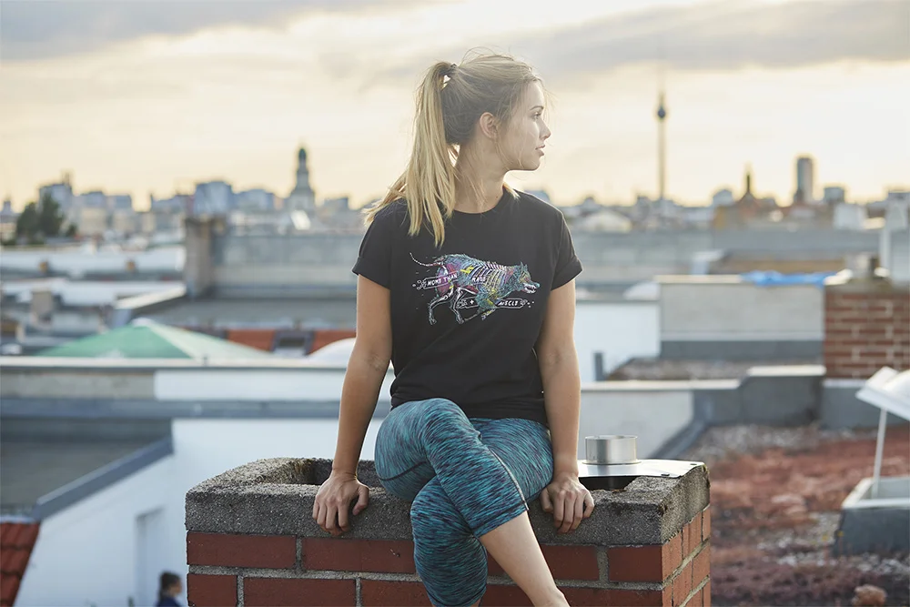 A young woman with blonde hair in a ponytail, wearing a black graphic t-shirt and colorful leggings, sitting on a brick rooftop ledge in an urban setting during sunset, with city skyline in the background.