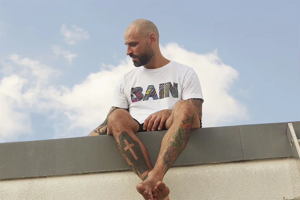 A bald man with a beard and tattoos sitting on a rooftop, looking down at his leg.