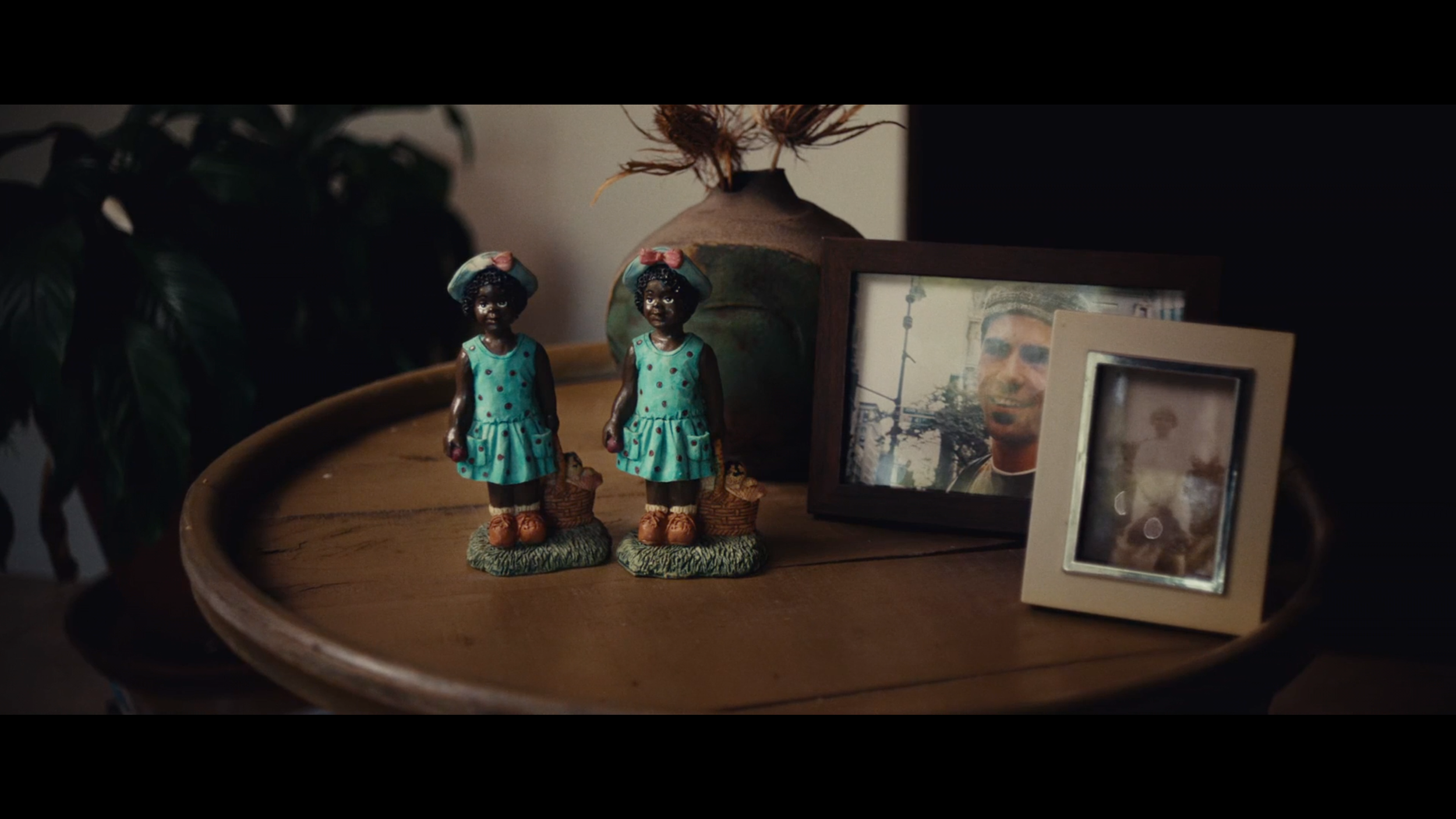 Decorative table with two black figurines of young girls wearing blue dresses and matching headbands, a large vase with dried plant, and framed photos of a man, all set on a wooden surface.