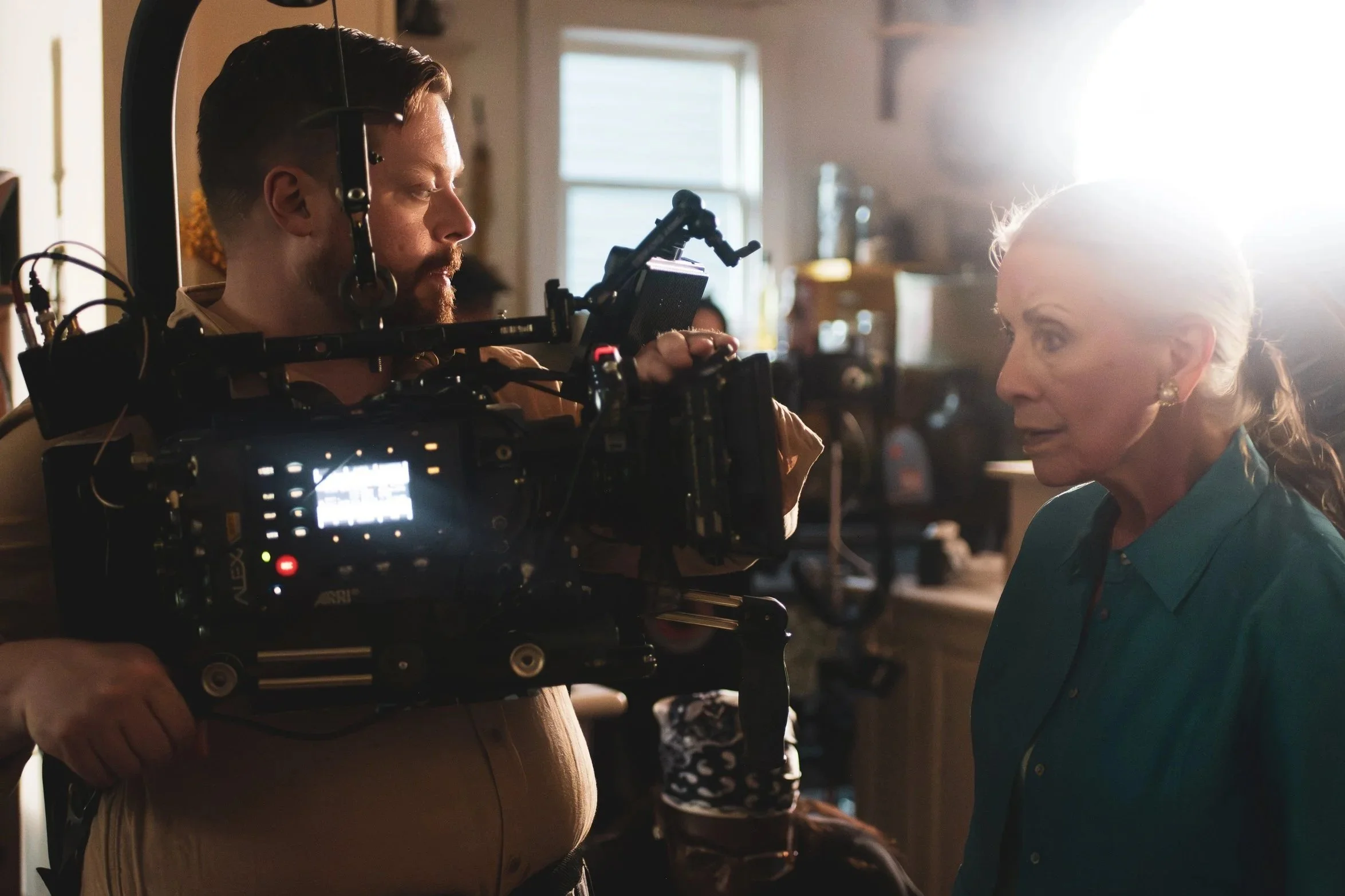 Behind-the-scenes shot of a film set showing a camera operator filming a woman with light-colored hair, dressed in teal, in a warmly lit indoor space.
