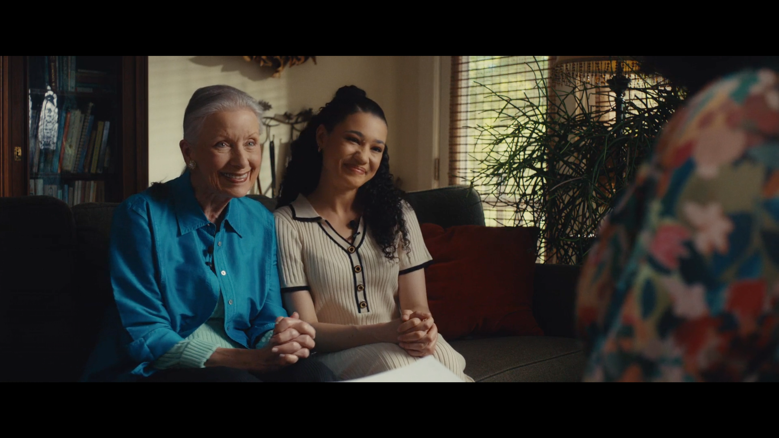Two women sitting on a couch smiling, one older with gray hair in a blue shirt, the other younger with dark curly hair in a striped cream and black dress, in a cozy living room with bookshelves, plants, and window blinds.