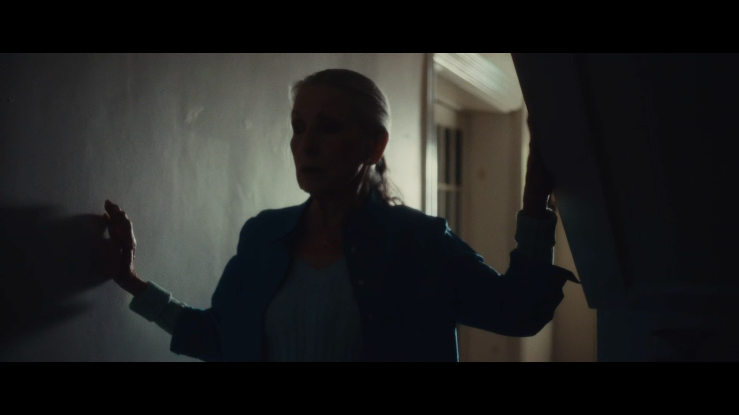 A woman with gray hair, wearing a blue jacket, stands in a dimly lit room with her hands on a wall, appearing distressed as she faces a person in a dark suit holding her wrists.
