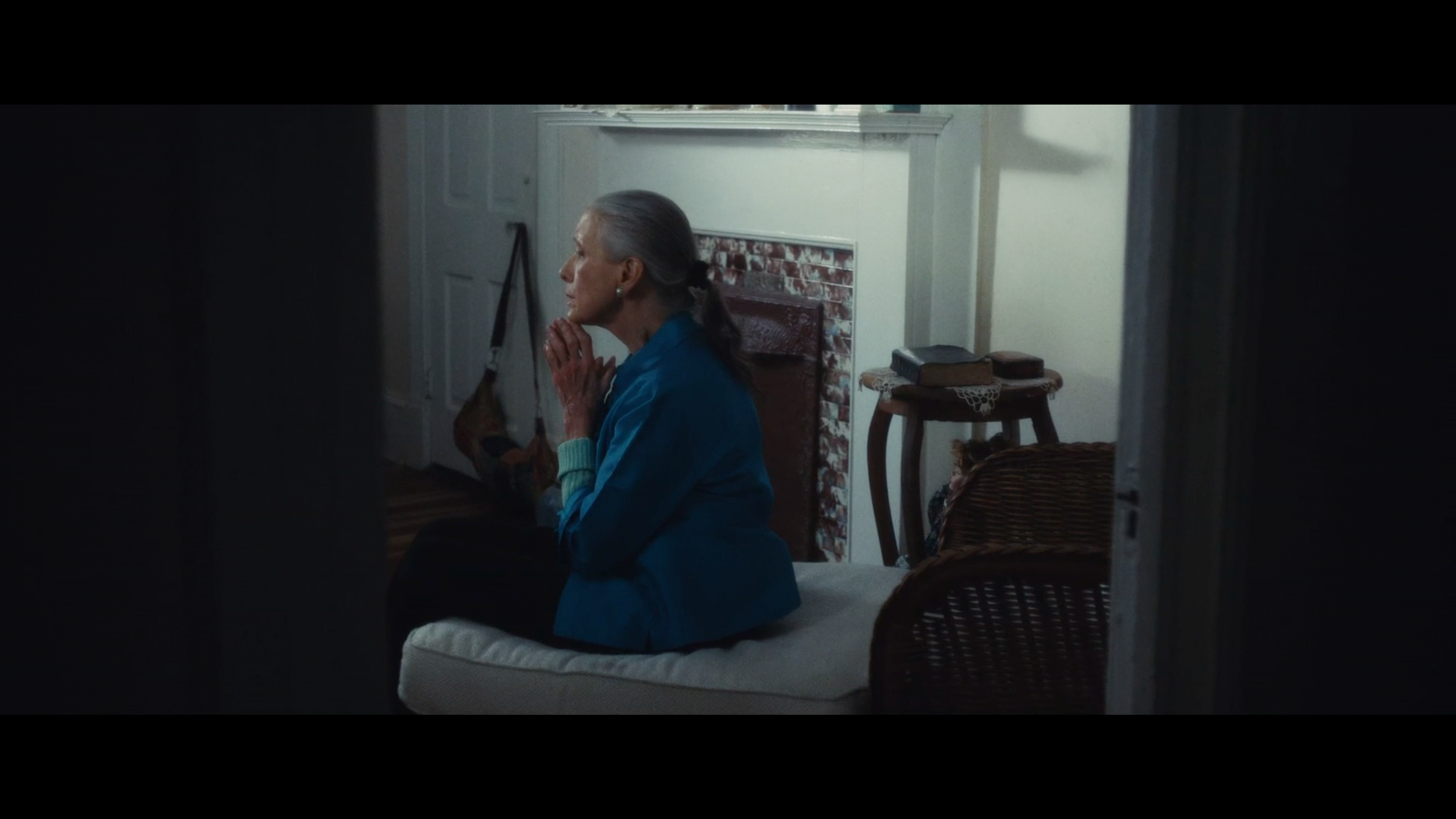An elderly woman with gray hair tied back, sitting on a bed with a contemplative expression, hands clasped near her face, in a cozy room with a vintage fireplace and furniture.
