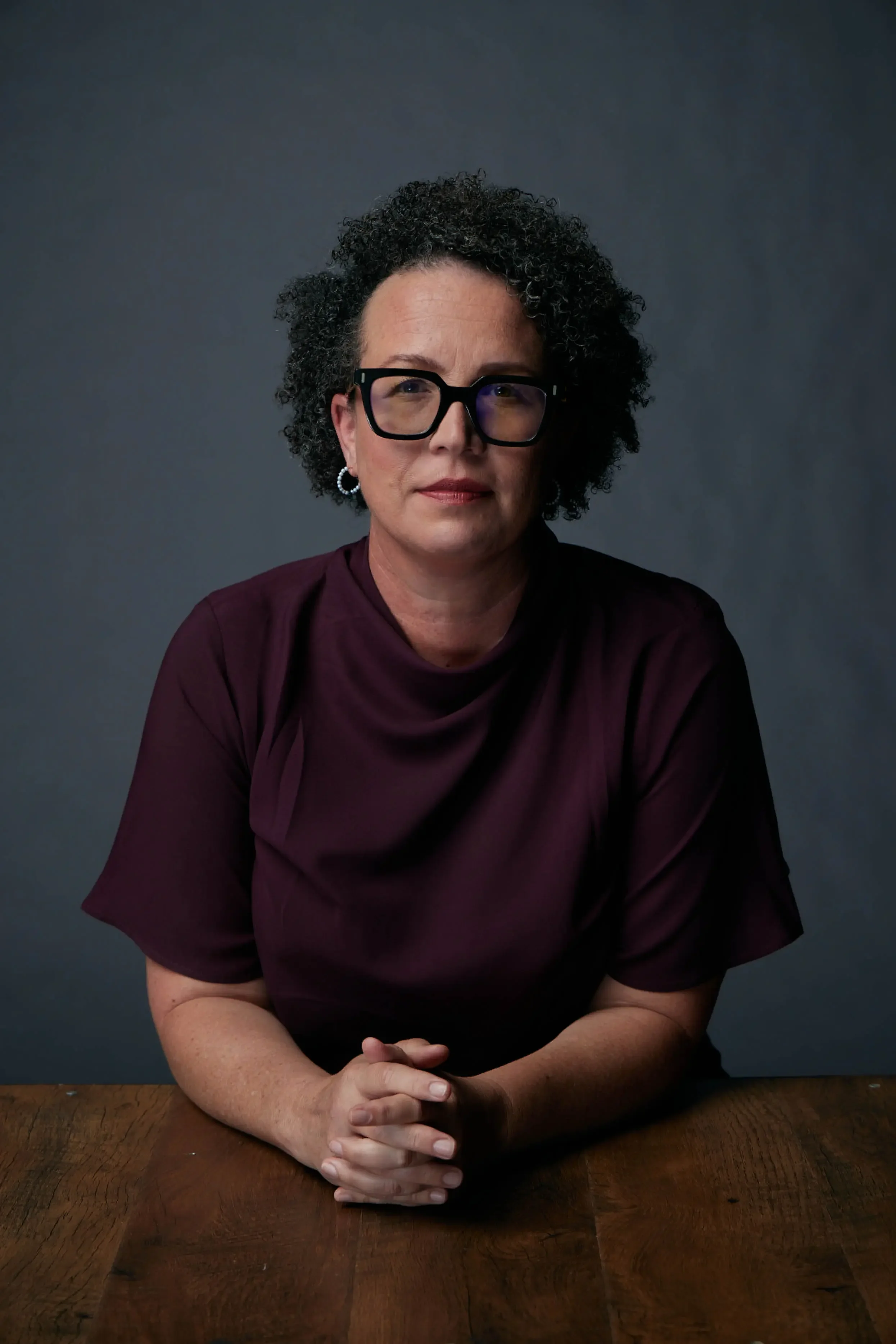 A woman with curly hair, wearing large black glasses and pearl earrings, sitting at a wooden table against a plain dark background, with her hands clasped on the table.