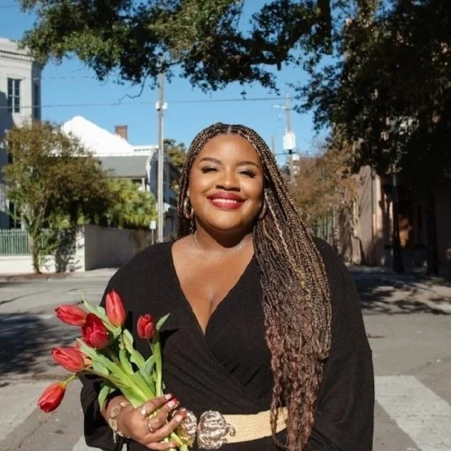 A woman holding a bouquet of red tulips, smiling outdoors on a sunny day with trees and buildings in the background.