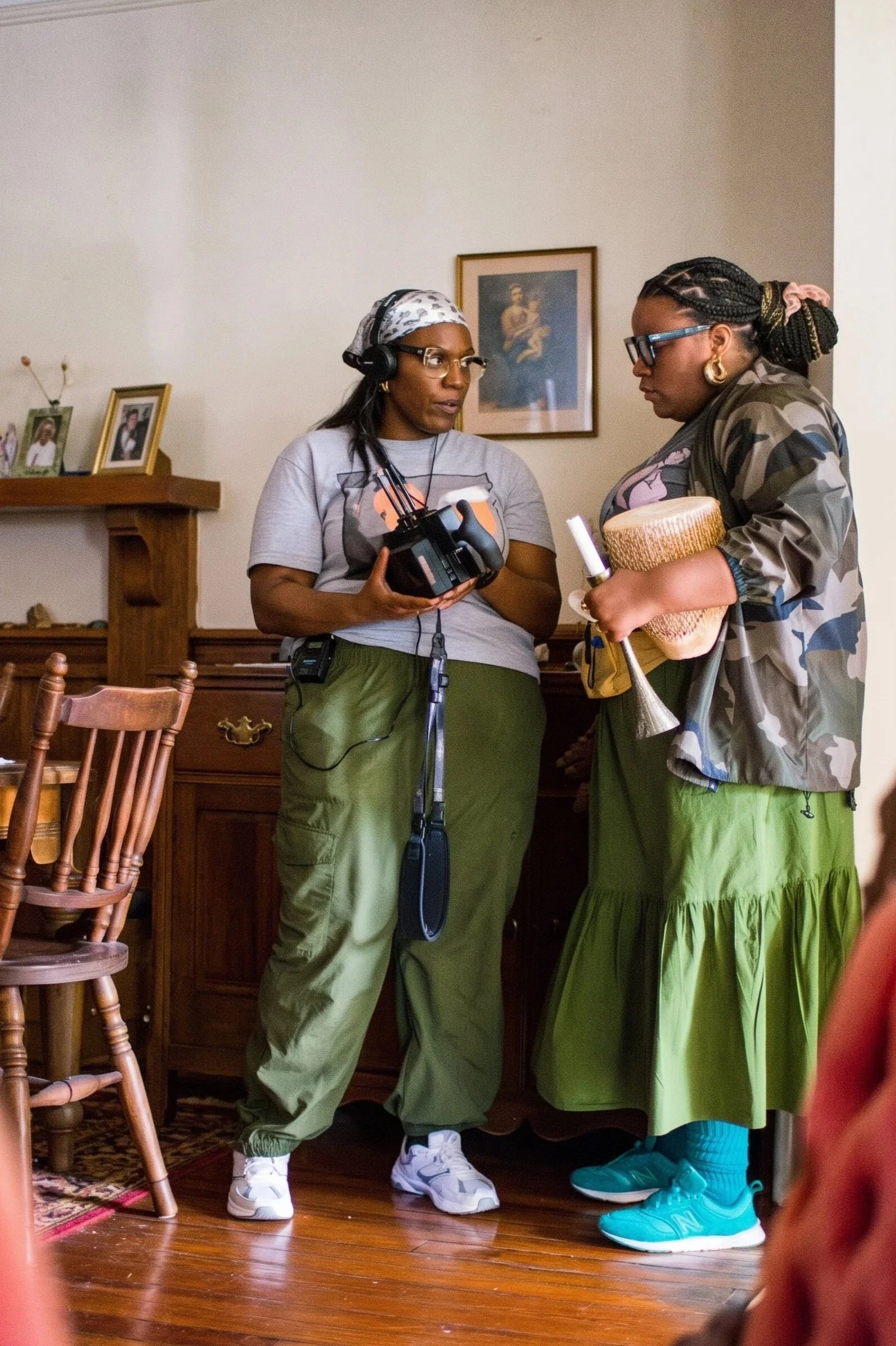 Two women are standing inside a room with wooden flooring and a dresser. One woman is holding a camera and wearing headphones, glasses, a headscarf, gray t-shirt, and green pants. The other woman is holding a basket with a wine bottle, wearing glasses, gold earrings, a camouflage jacket, a green skirt, and teal sneakers. There are framed photographs and a painting hanging on the wall behind them.