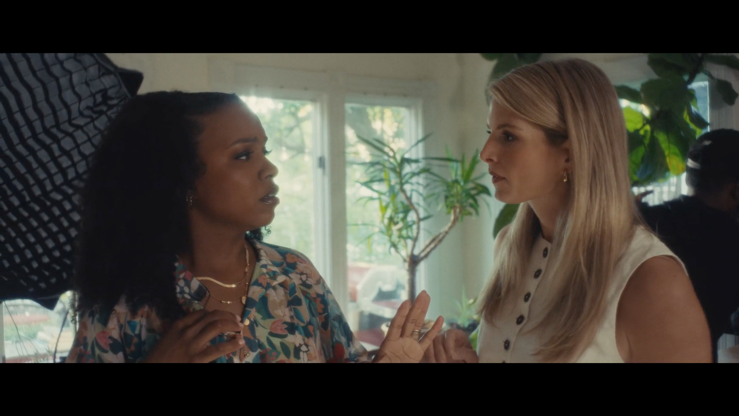 Two women engaged in a conversation inside a room with large windows and plants, one woman with dark curly hair wearing a patterned top, the other with light blonde hair in a sleeveless blouse.