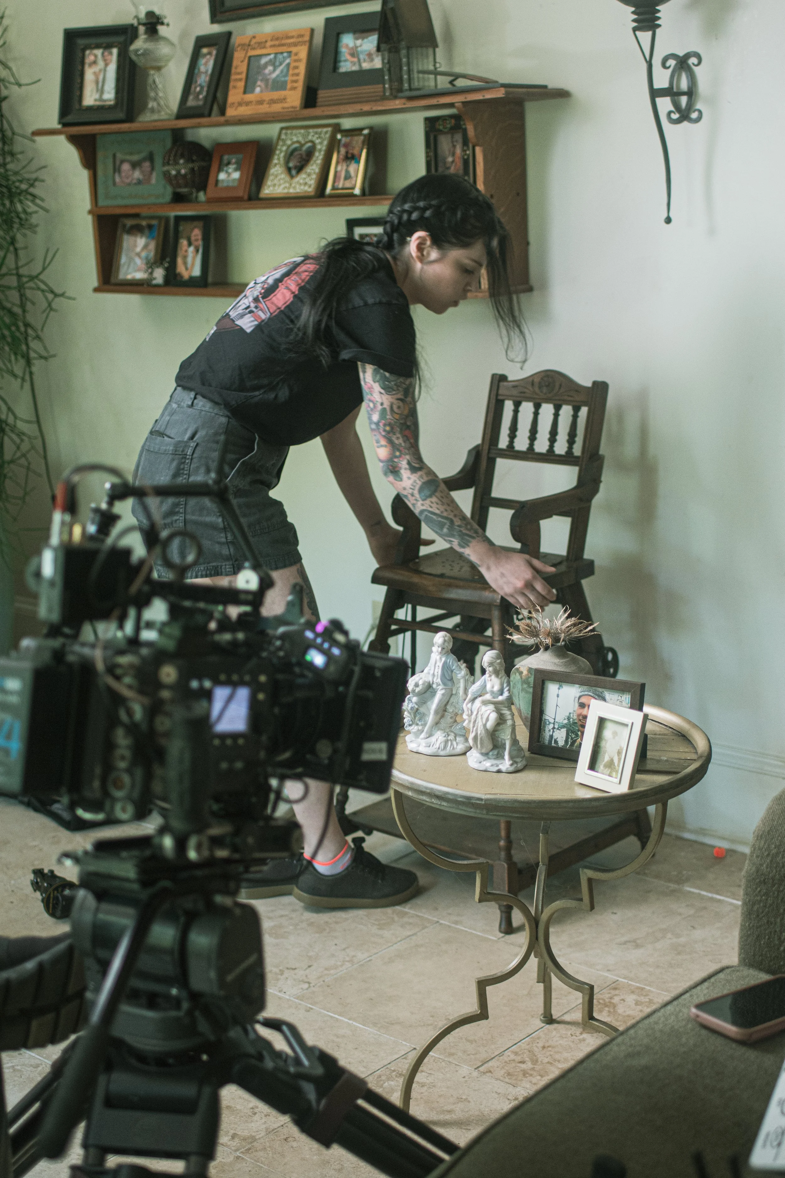 A woman with dark hair, tattoos on her arms, and wearing casual clothes is adjusting a wooden chair in a room with decorative items, framed photographs, and various figurines on a table. A professional camera is positioned nearby, capturing the scene