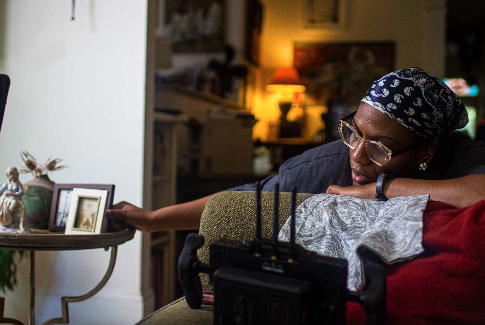 A woman wearing glasses and a blue patterned headscarf sits on a couch in a cozy living room, resting her head on a pillow with her eyes closed. In the background, a lamp emits warm light, and the room is decorated with various framed pictures, a sculpture on a side table, and plants.