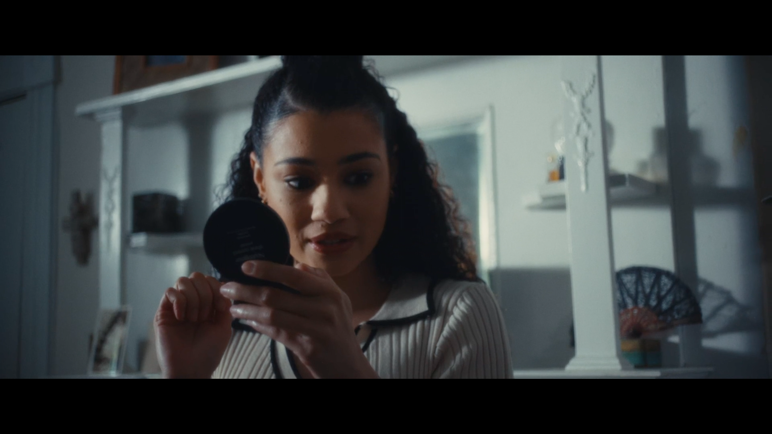 A young woman with curly hair and a top knot looking into a mirror and holding a makeup compact.