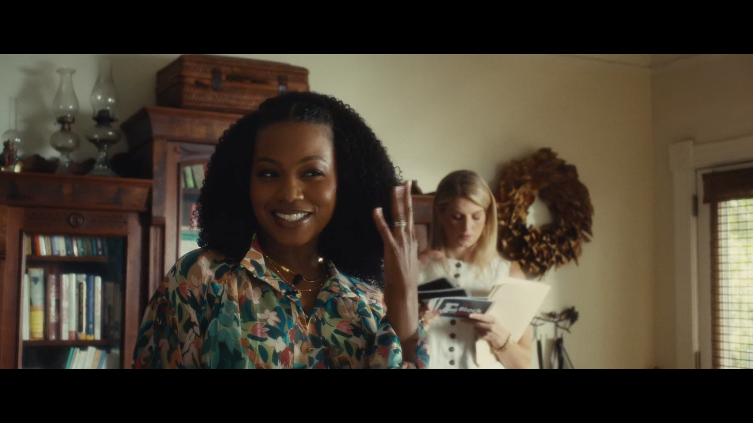 Smiling woman with dark curly hair wearing a colorful floral shirt, waving with her right hand, in a room with wooden furniture and a bookshelf, with another woman in the background reading a magazine.