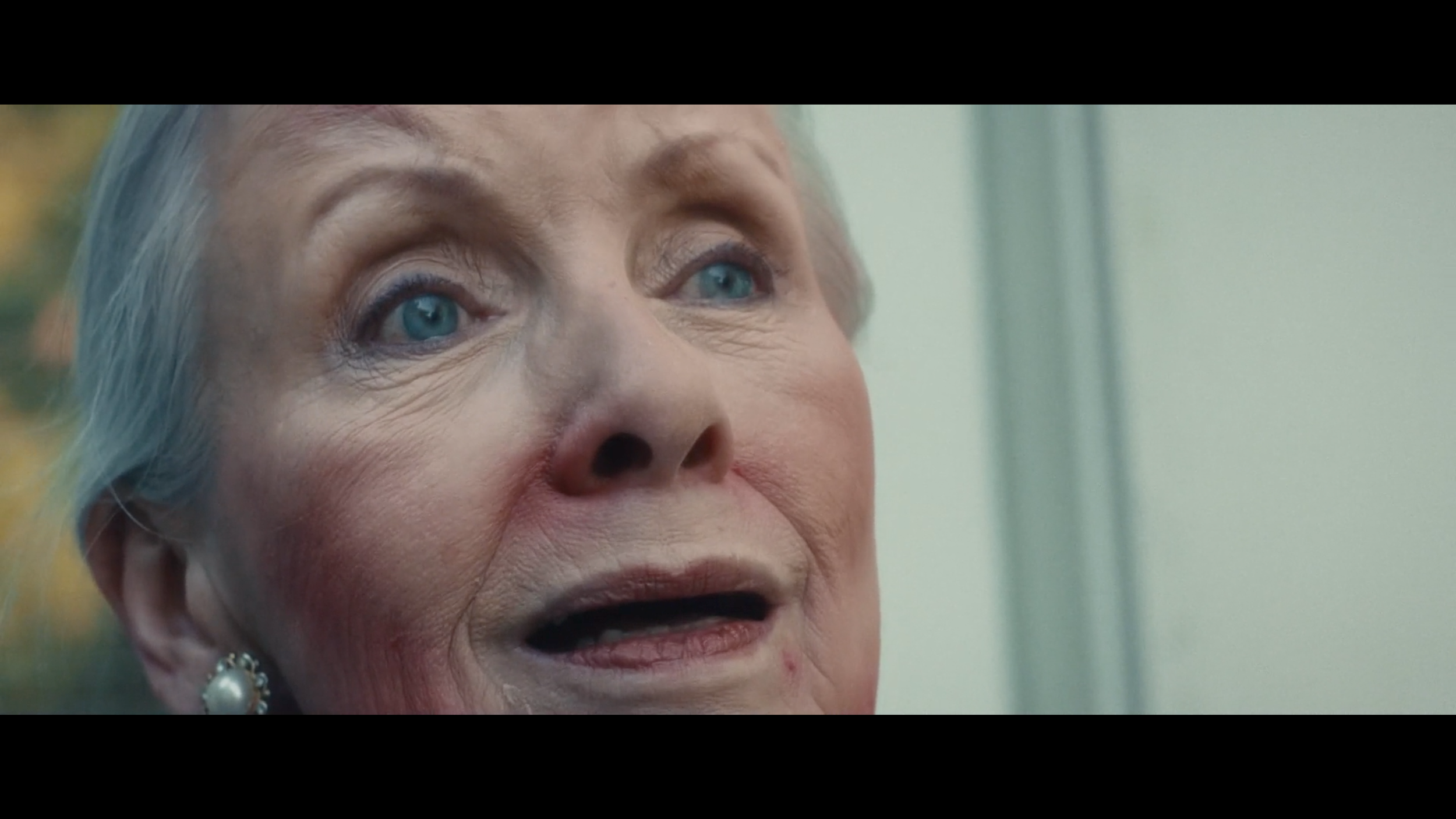 Close-up of an elderly woman's face with gray hair and blue eyes, wearing a pearl earring. She appears to be speaking or reacting emotionally.