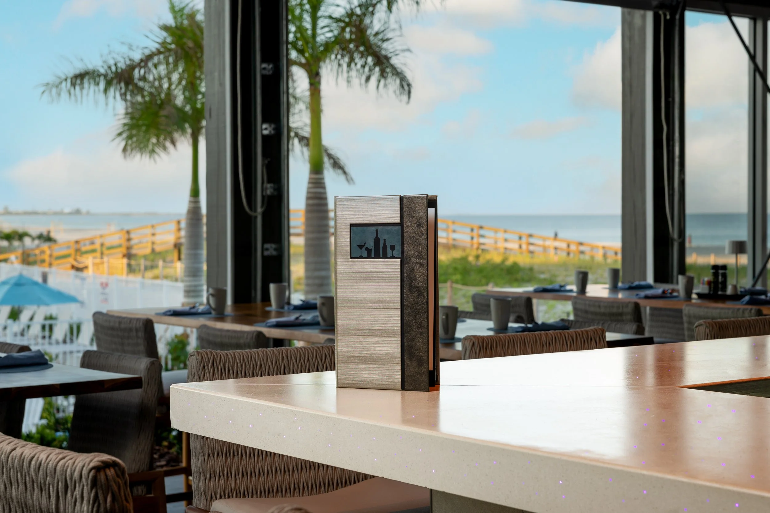 Indoor dining area with a view of the beach and palm trees through large windows, table settings with cups and napkins, and a menu stand on a white countertop.