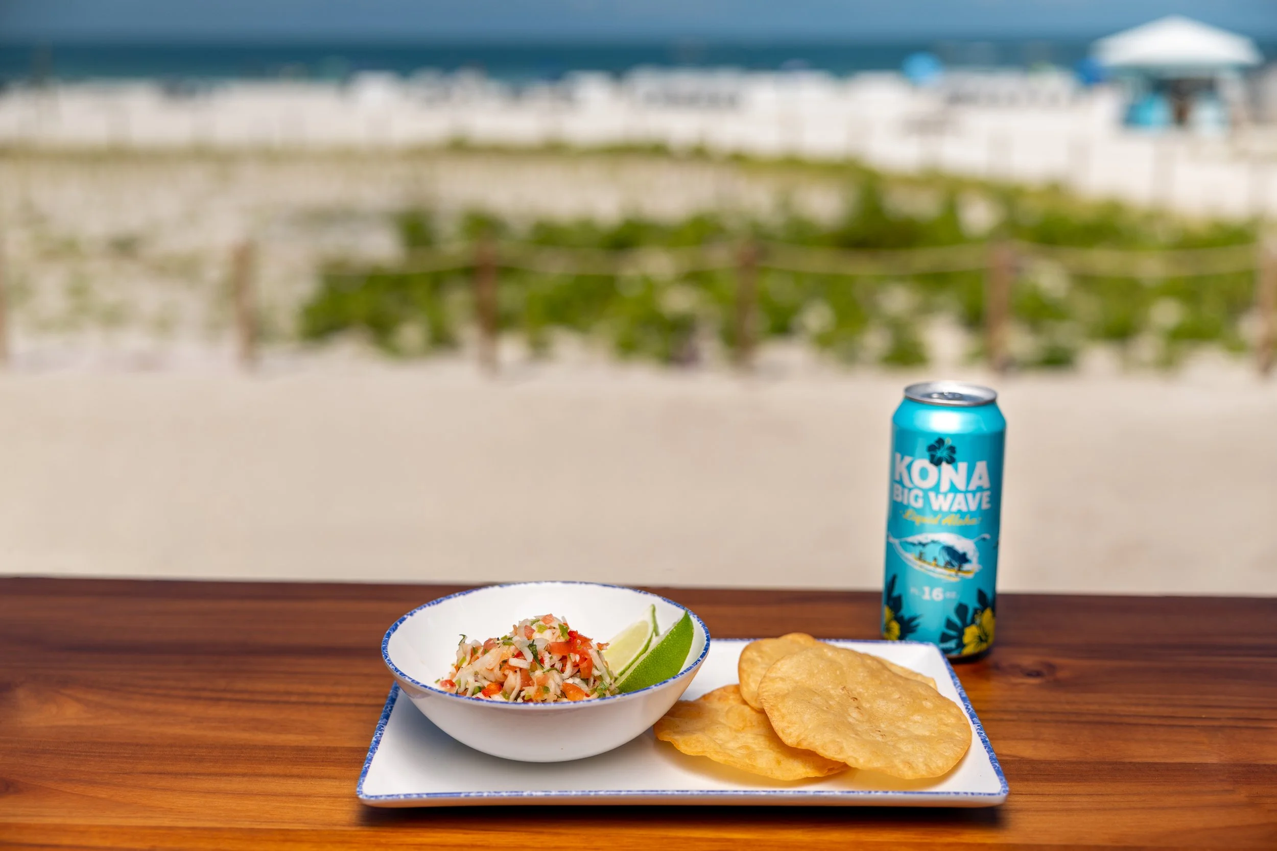 A plate of fish ceviche with lime wedges and salted crackers on a wooden table, with a blurred beach and ocean in the background, featuring white sand, green plants, and beach umbrellas.
