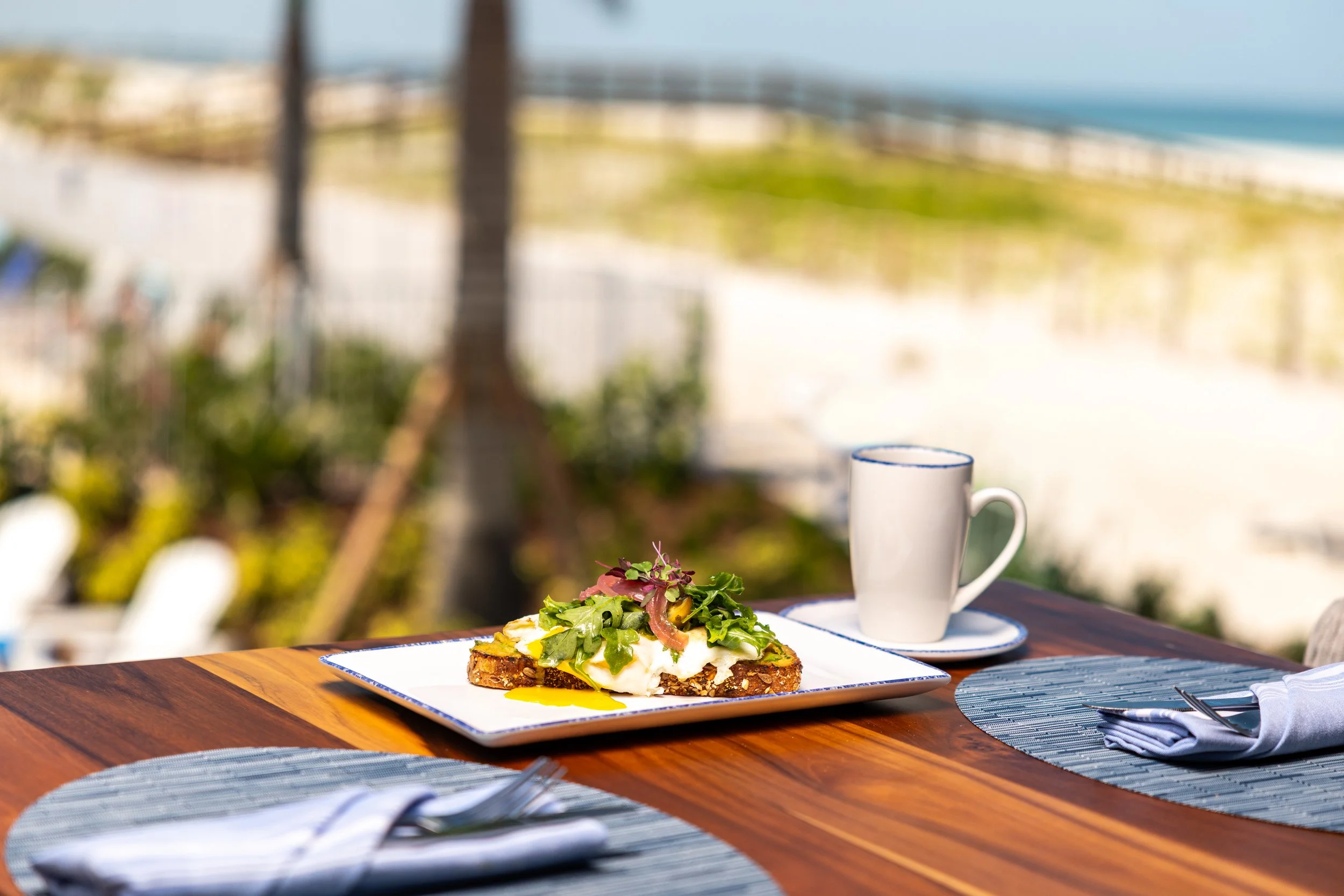 An outdoor dining table with a plate of avocado toast topped with greens, onions, and microgreens, a white coffee mug, and blue placemats, overlooking a tropical beach scene.