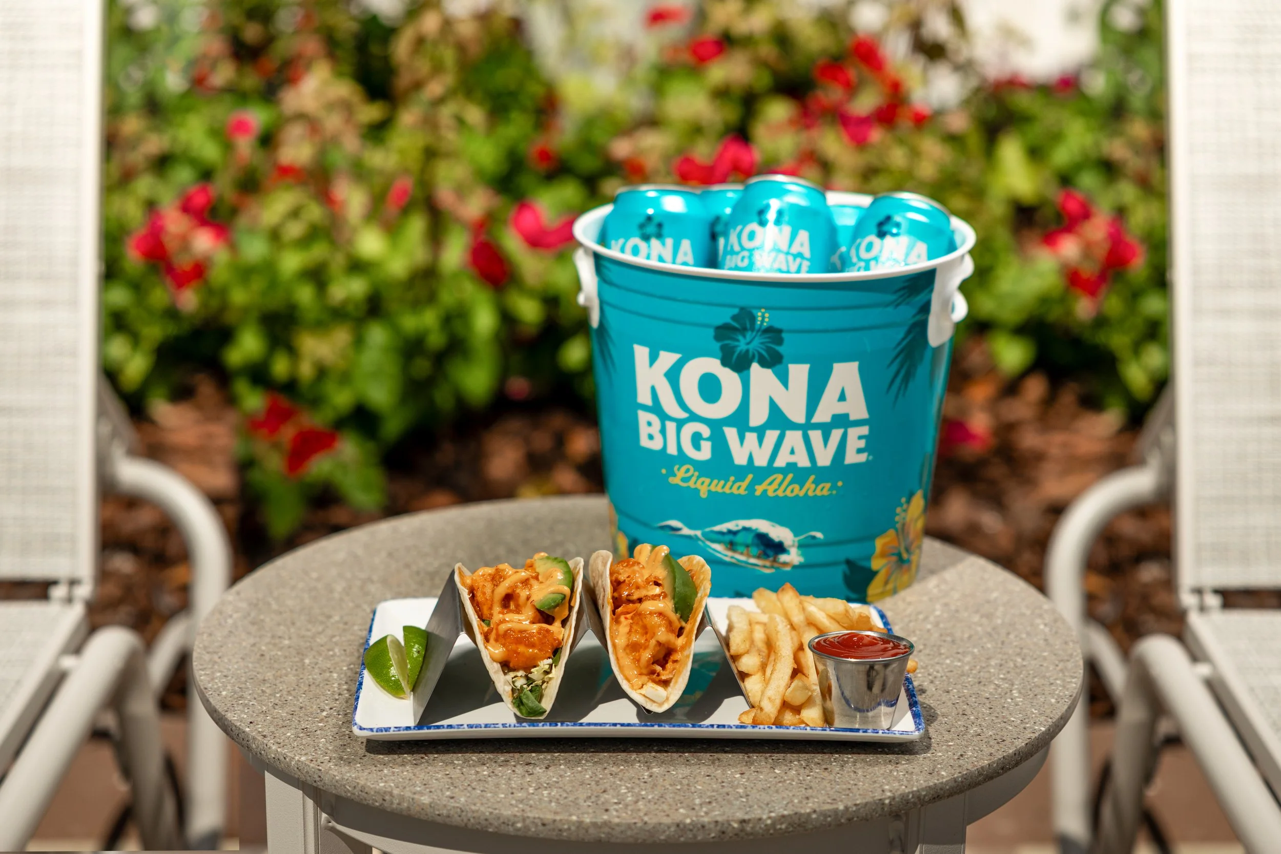 A table with a tray of three soft tacos filled with meat, cheese, and vegetables, a container of French fries, and a small cup of ketchup, with a tropical-themed bucket of Kona Big Wave beer and some lime wedges on a patio outside, with greenery and 