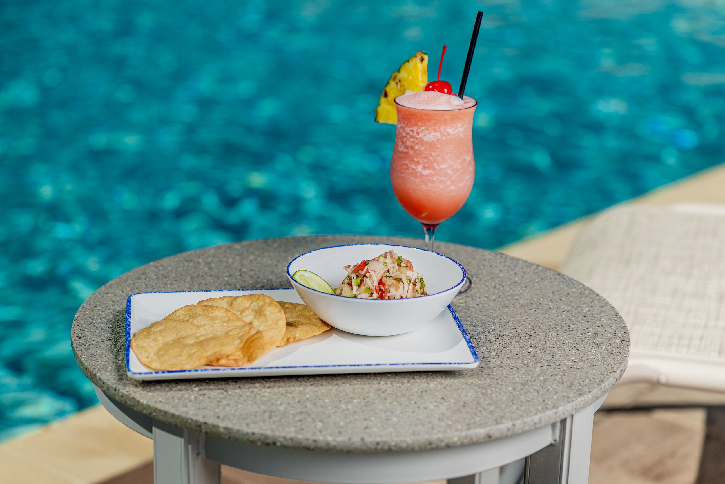 A tropical drink with a cherry and pineapple garnish on a table next to a plate of fried food and a seafood salad near a swimming pool.