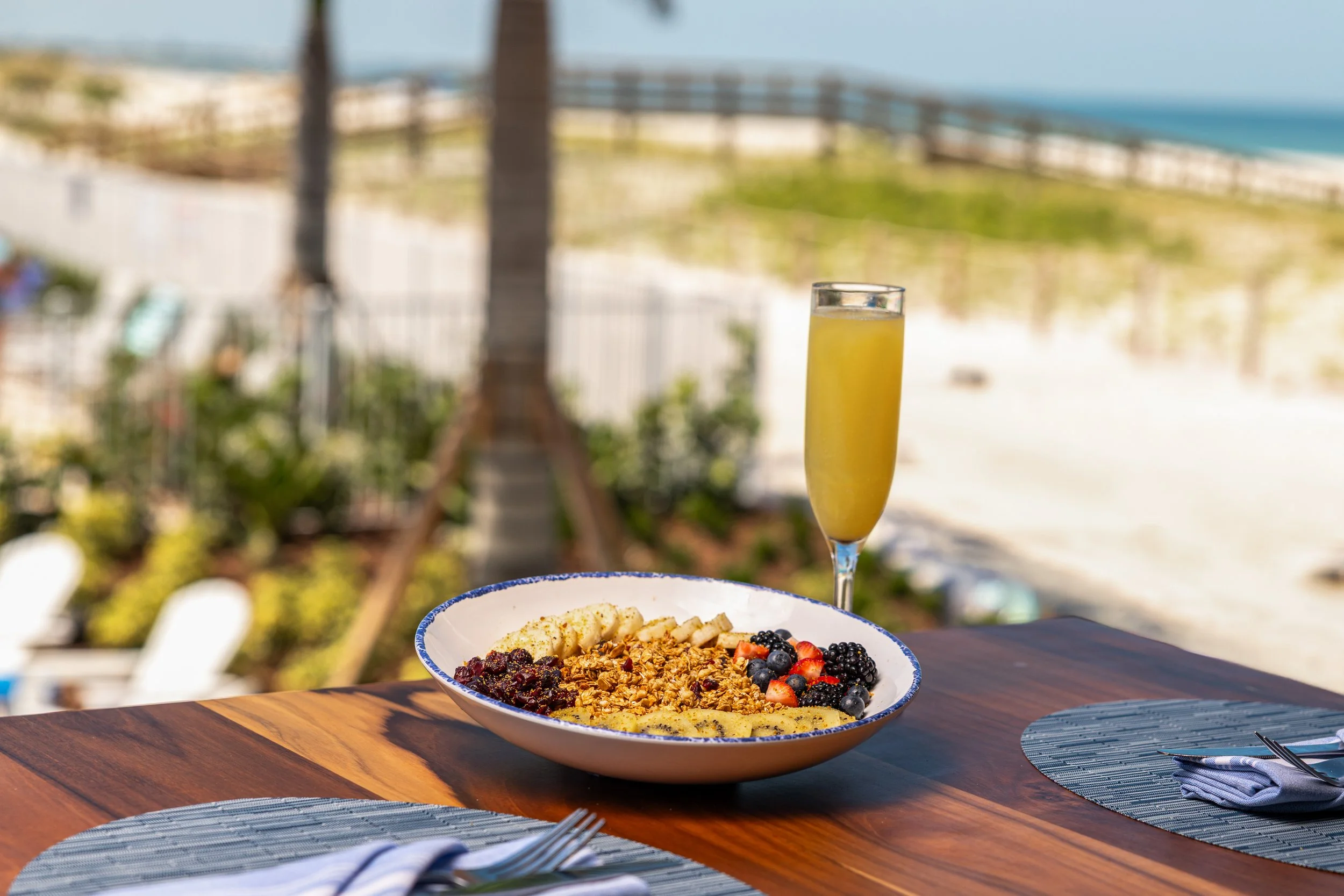 A bowl of mixed berries, granola, and sliced fruit on a dining table outdoors with ocean and palm trees in the background, along with a glass of yellow beverage.