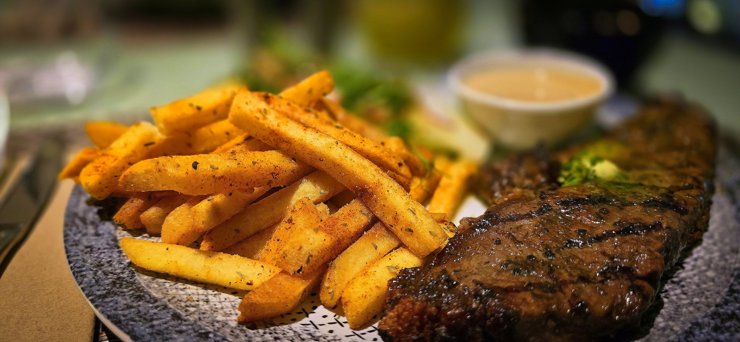 French fries seasoned with herbs, a grilled steak with green garnish, and a small bowl of dipping sauce, all on a restaurant plate.