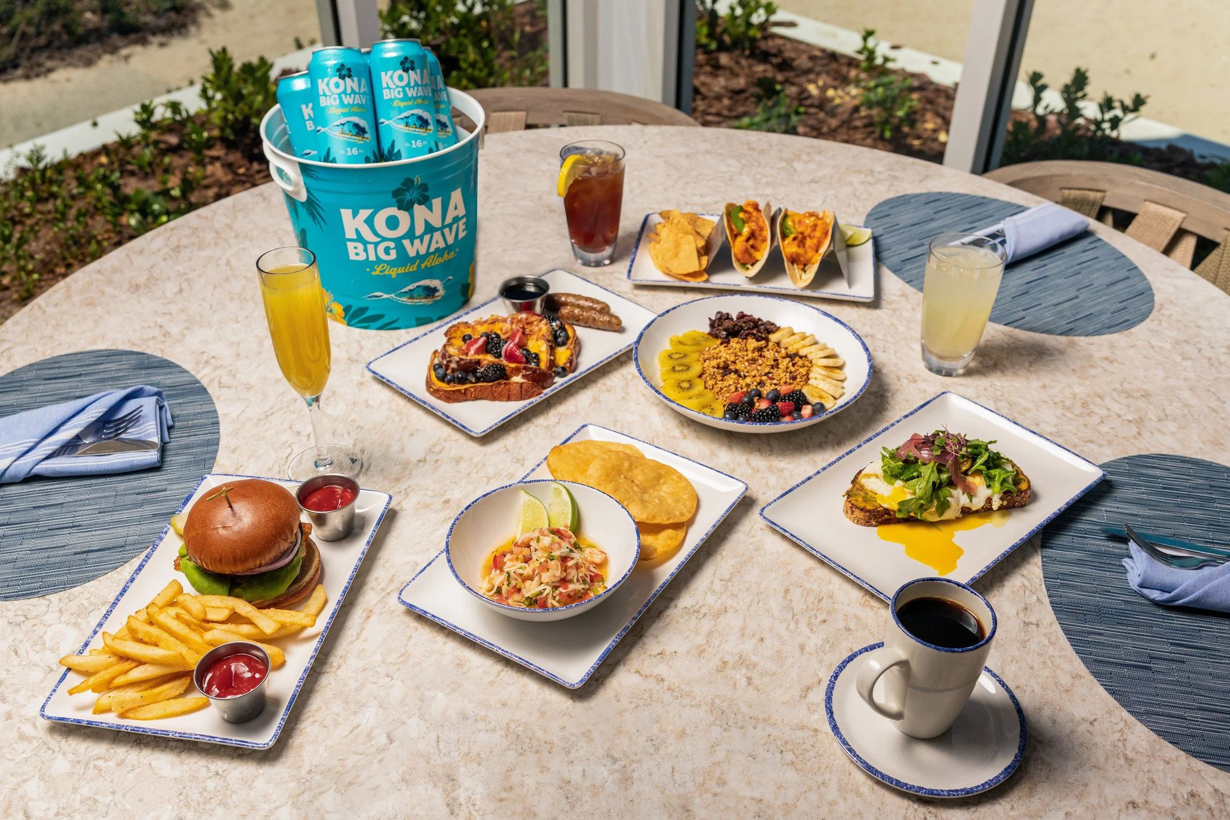 A round outdoor table set with various dishes, including a burger, fries, fruit-topped toast, tacos, small bowls of sauces, a salad, and drinks, with a large blue bucket labeled 'Kona Big Wave' beer and a glass of iced tea with lemon.