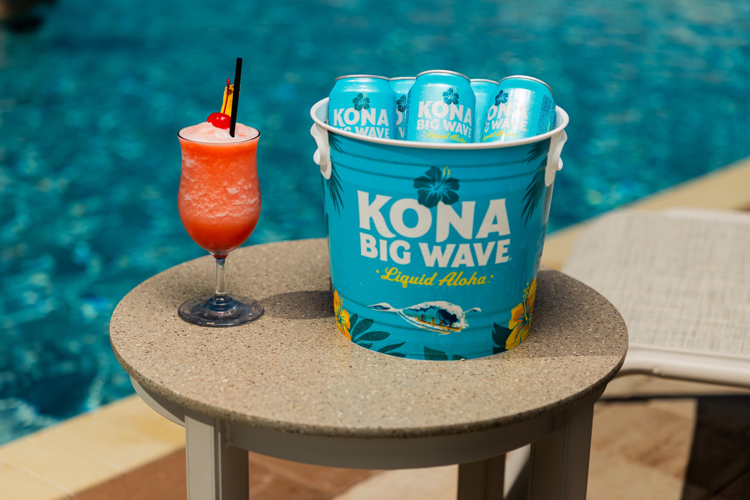 A tropical drink with a cherry, a yellow flag, and a black straw next to a bucket of Kona Big Wave beer cans by a swimming pool.