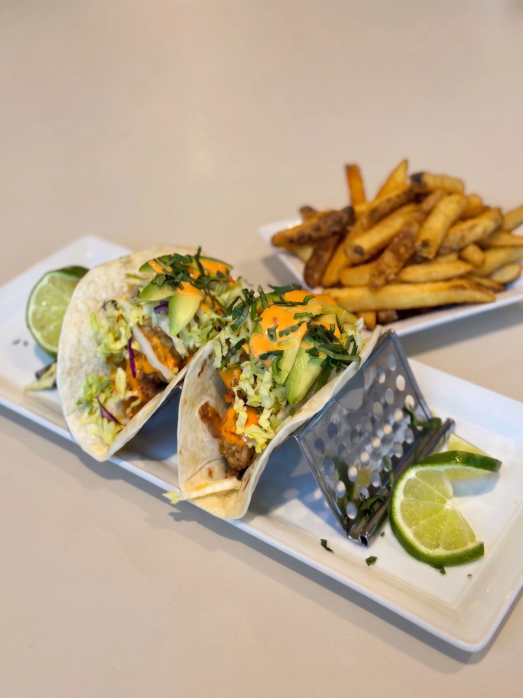 Three fish tacos topped with shredded lettuce, sliced cucumber, and chopped herbs, served with a side of French fries and lime wedges on a white rectangular plate.