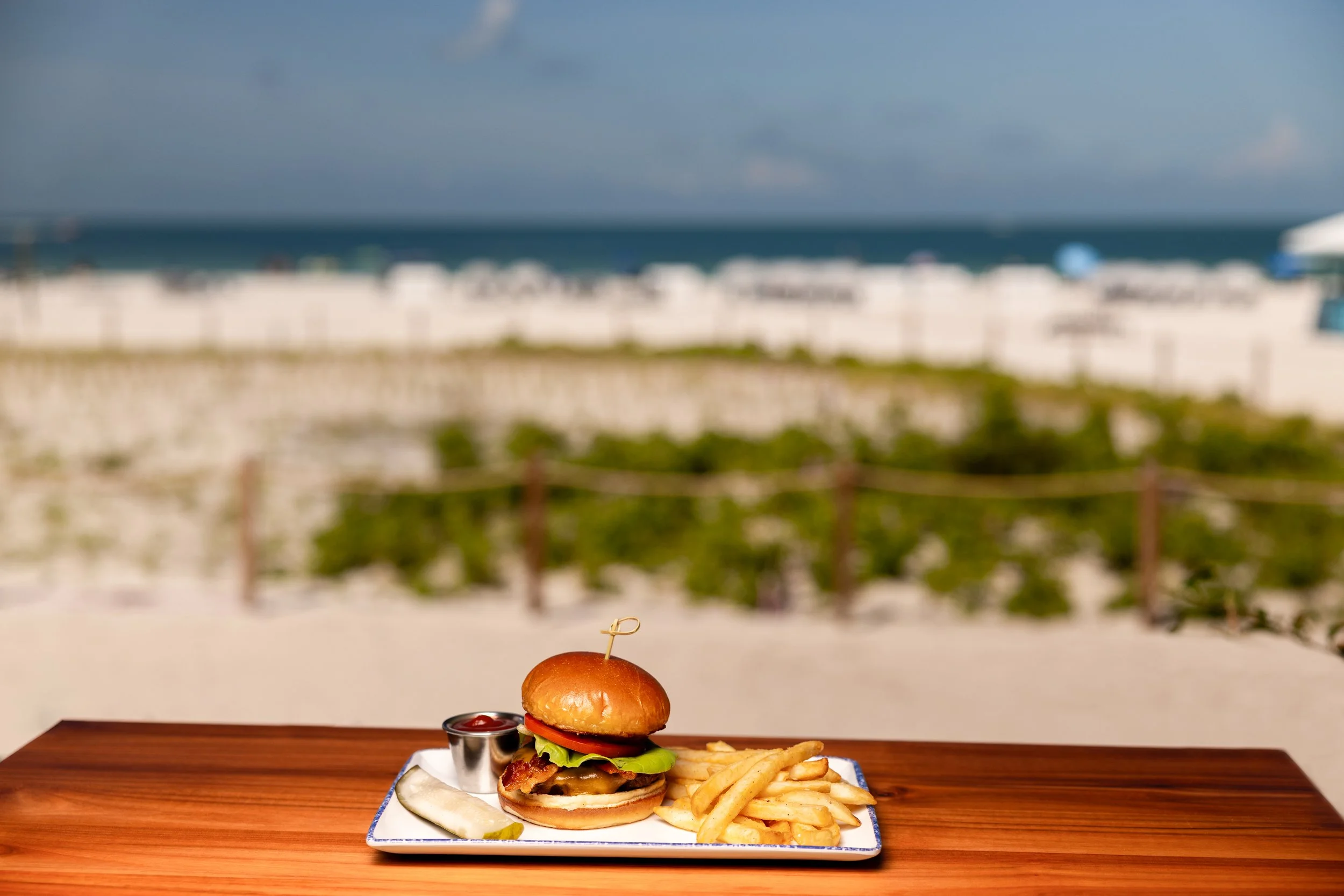 A cheeseburger with lettuce, tomato, and bacon, served with fries and ketchup on a white plate, on a wooden table outdoors with a blurred ocean and sky background.