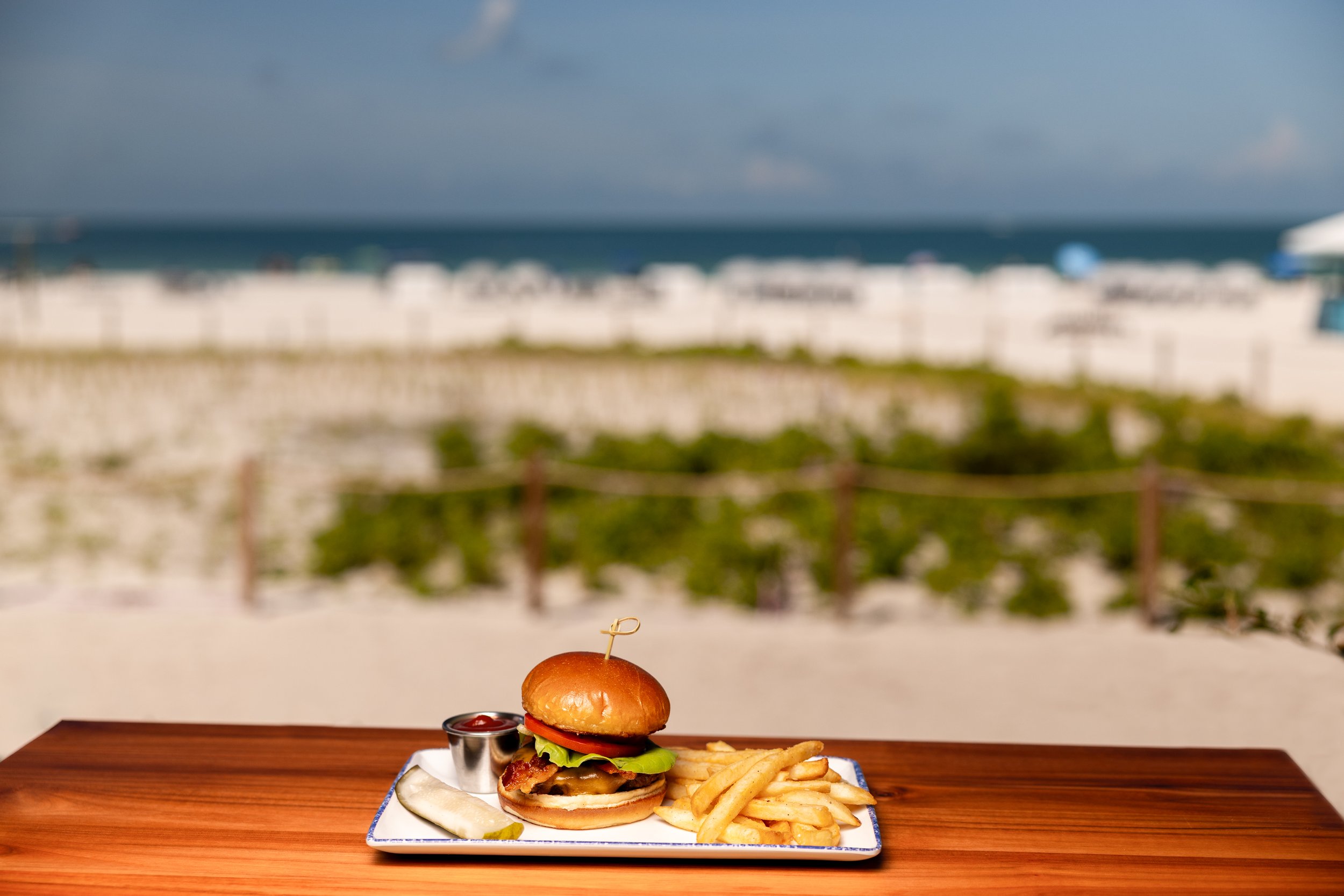 A cheeseburger with lettuce, tomato, melted cheese, and bacon, served with French fries and ketchup on a plate, on a wooden table indoors with a blurred seaside view in the background.
