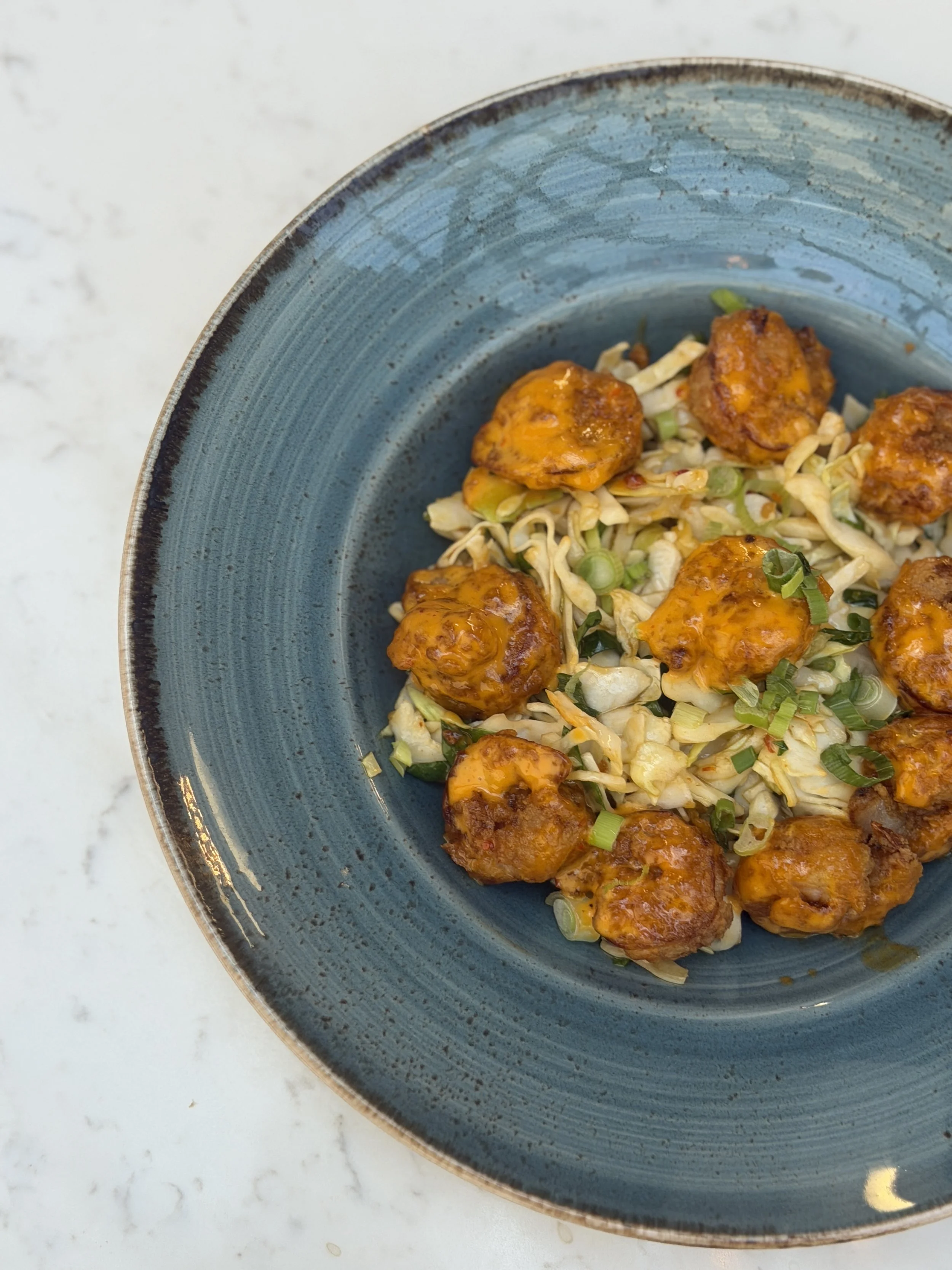 A blue bowl containing fried chicken pieces coated in sauce, garnished with chopped green onions, on a white marble surface.