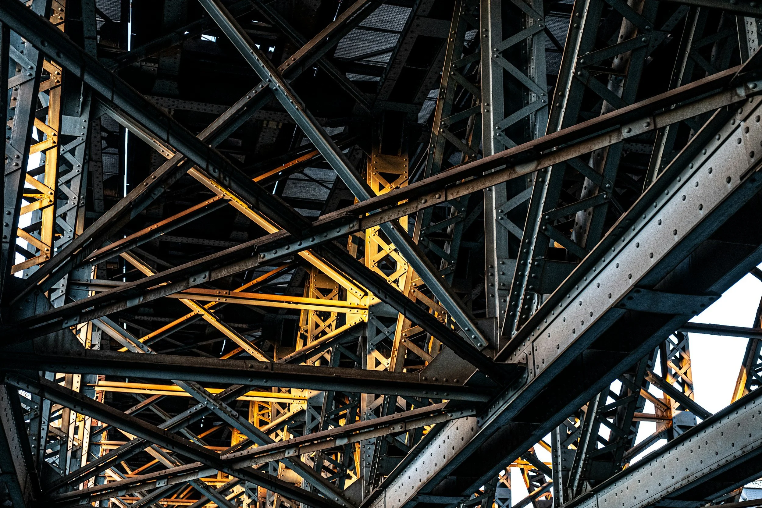 Close-up view of the metal framework of a large steel bridge, with crisscrossing beams and rivets, illuminated by lights.