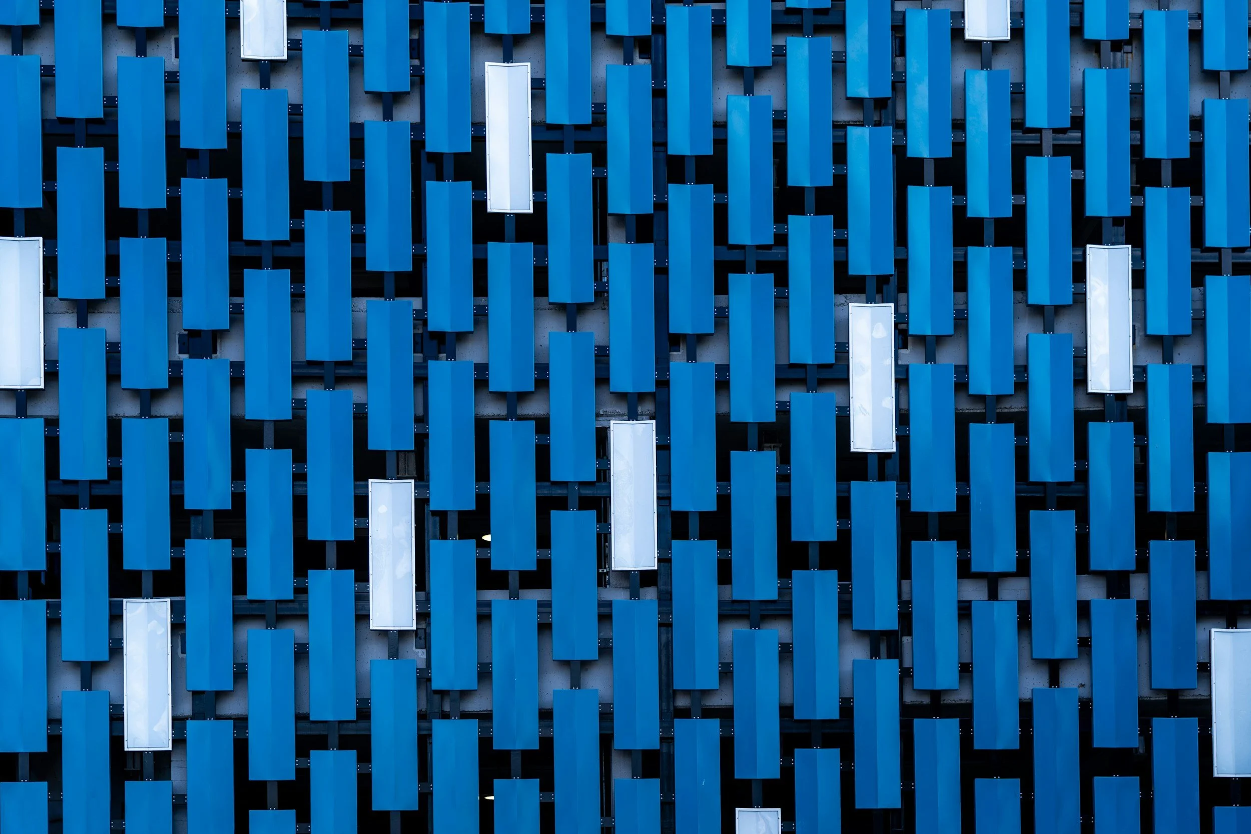 Close-up of a modern building facade with vertical blue rectangular panels arranged in a grid pattern, interspersed with a few white panels, creating a geometric abstract design.