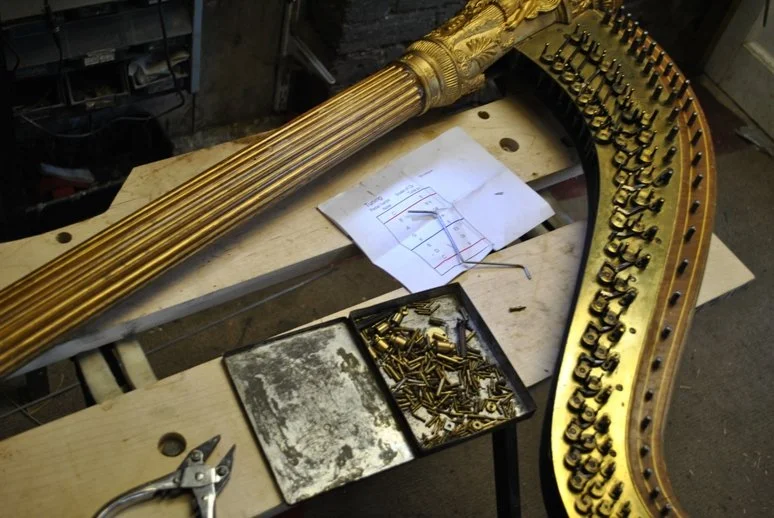 A workbench with a Grecian neck and pillar, partly assembled, gold in color, with tuning pegs and hardware. There are tools, a small tray of screws, and a diagram or plans on the work surface.