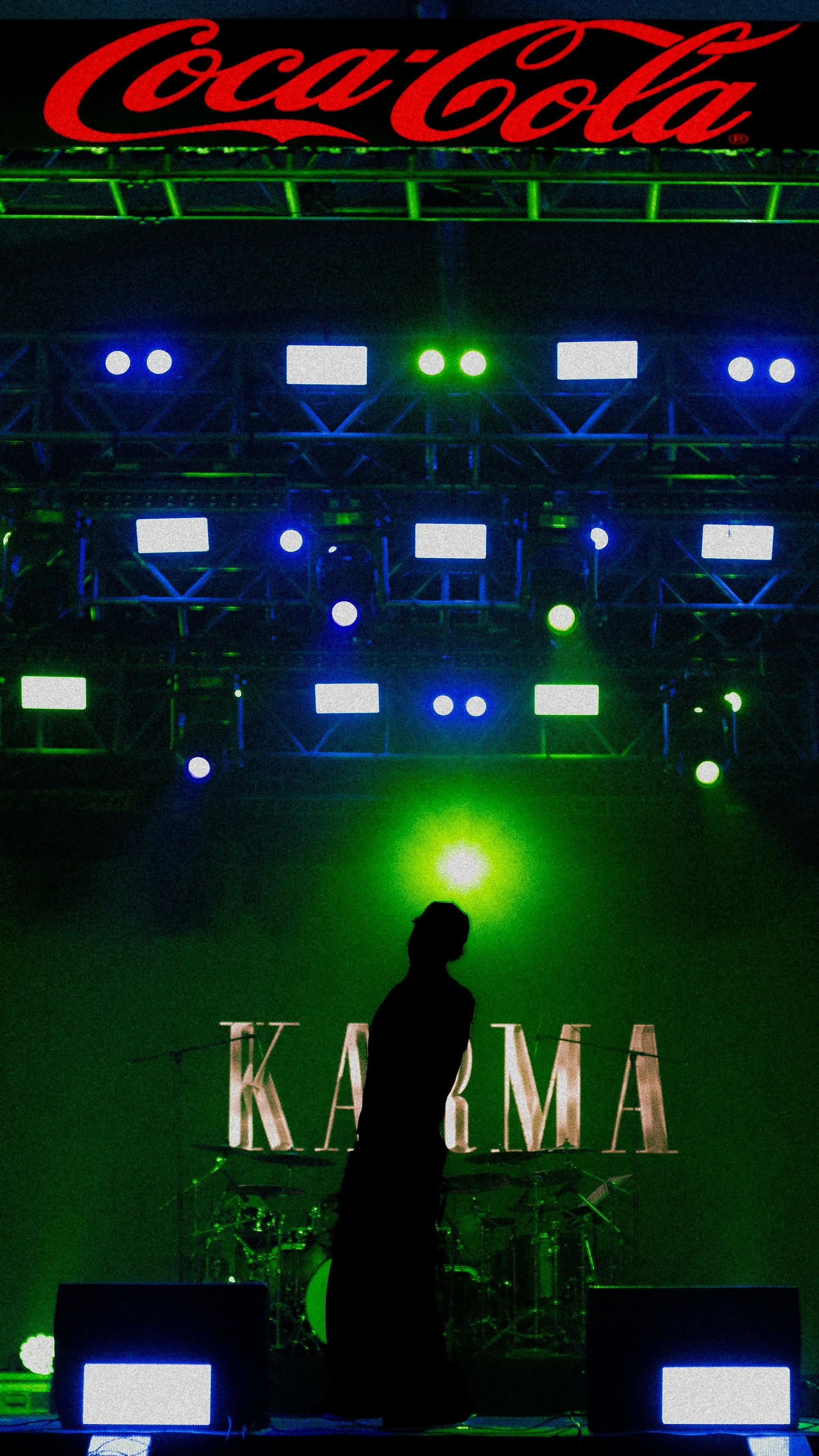 A silhouette of a person standing in front of a concert stage with green and blue lights, a large Coca-Cola sign above, and the word 'KARMA' displayed on the stage backdrop.
