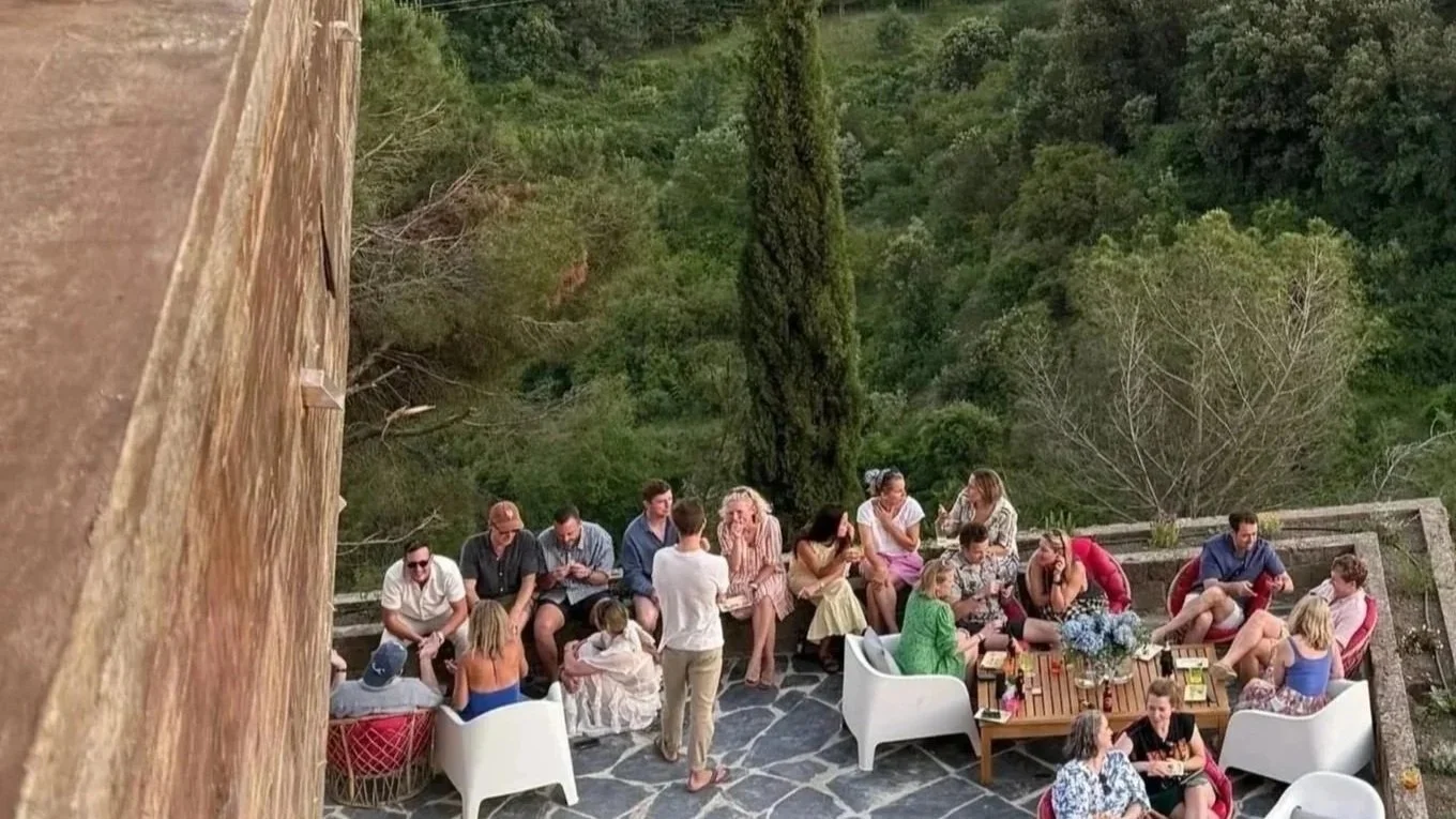 A group of people socializing on an outdoor terrace with lush green trees and a tall cypress tree in the background, some sitting on chairs and others standing or engaging in conversations.