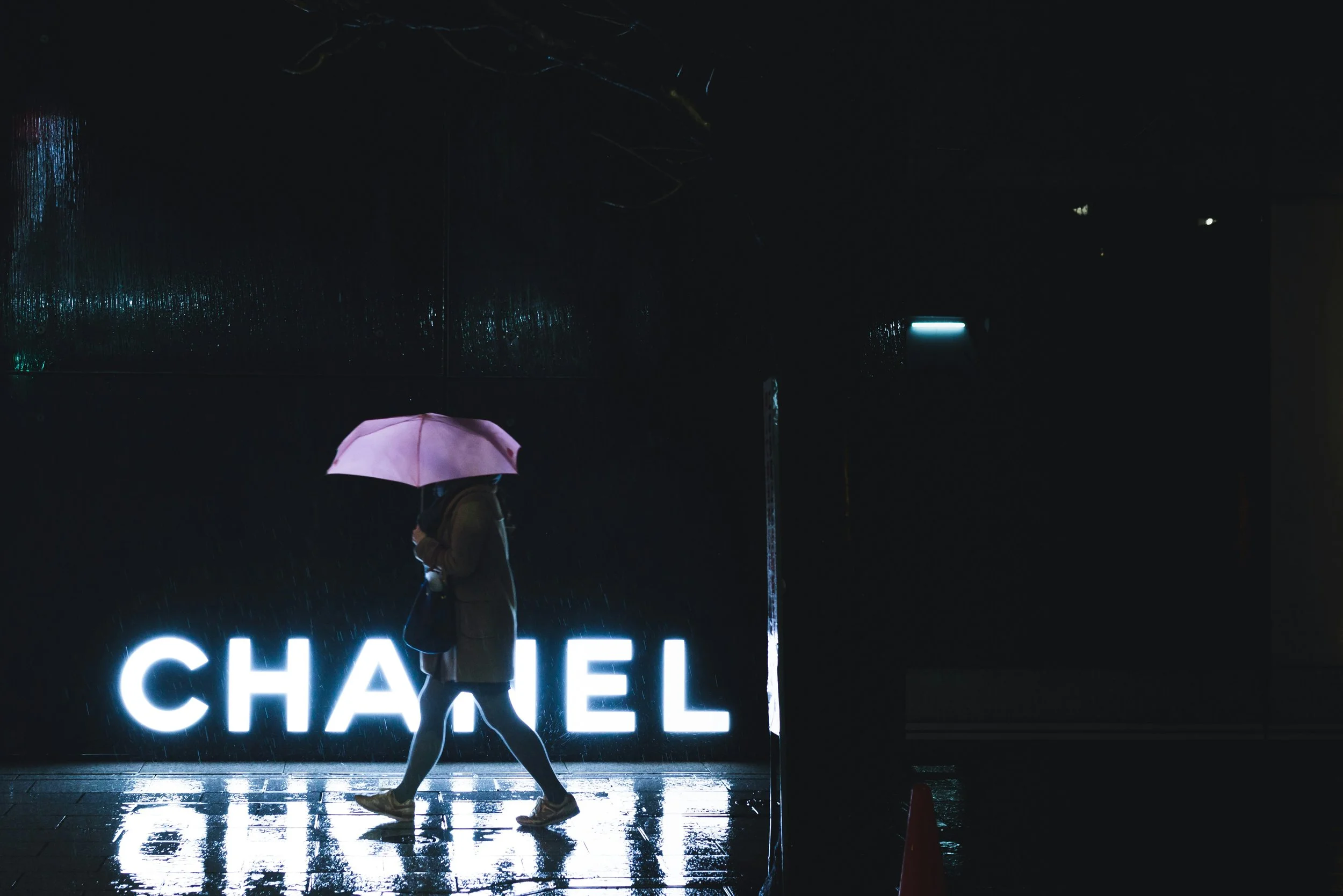 A person walking with an umbrella on a rainy night in front of a brightly lit Chanel store sign.