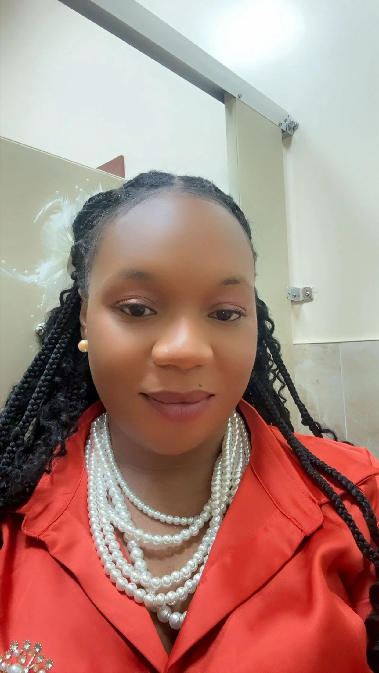 A woman wearing multiple strands of pearl necklaces and earrings, dressed in a red outfit, standing in front of a bathroom stall door.