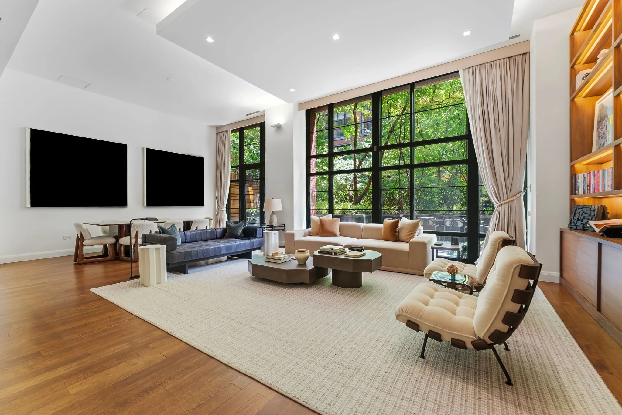 A spacious modern living room with large windows showing greenery outside, featuring a white sofa, beige chairs, a coffee table with books and decorative items, a textured rug, bookshelf, and wall-mounted televisions.