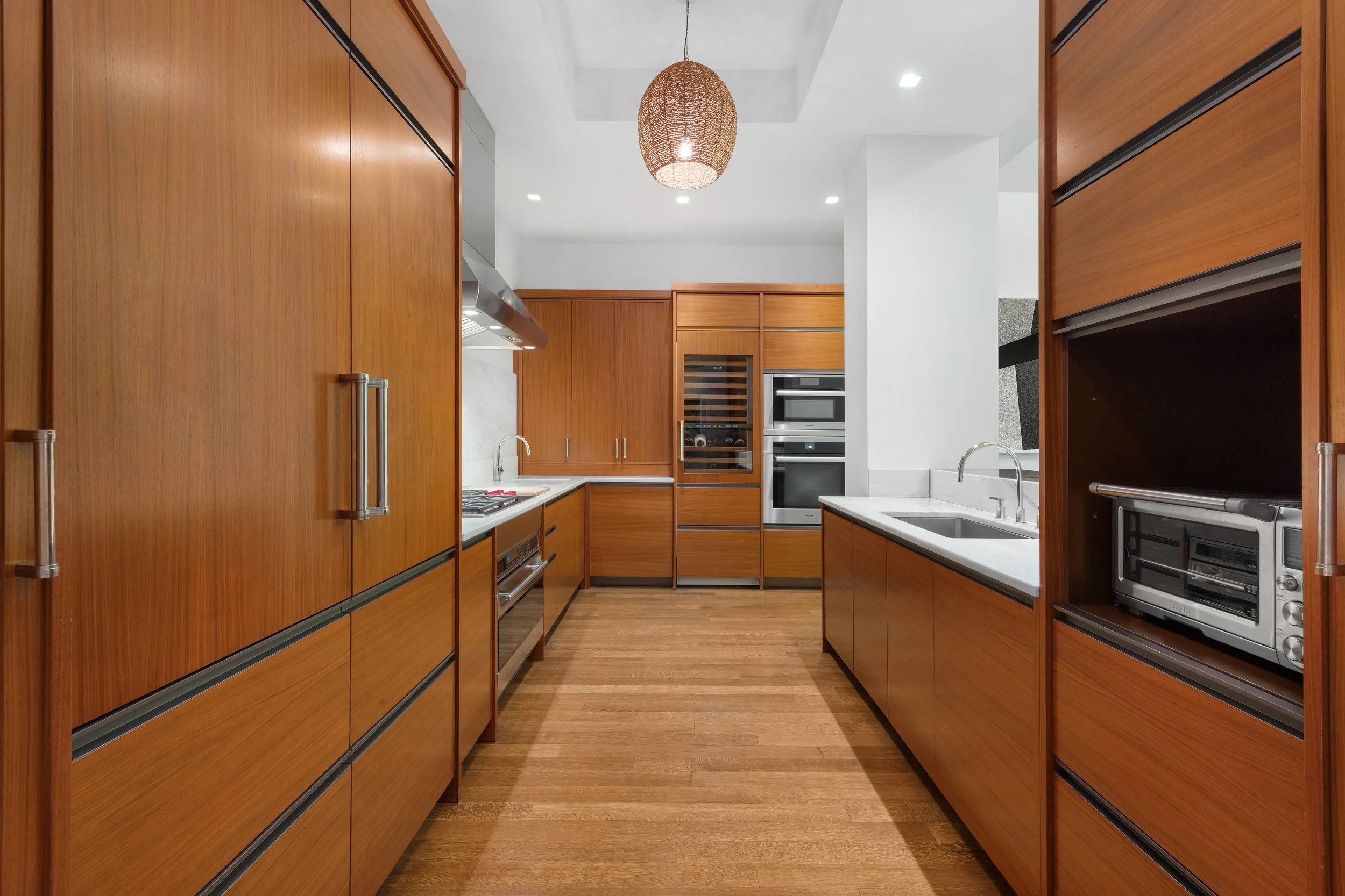 A modern kitchen with wooden cabinets, white countertops, built-in appliances, and a woven pendant light hanging from the ceiling.